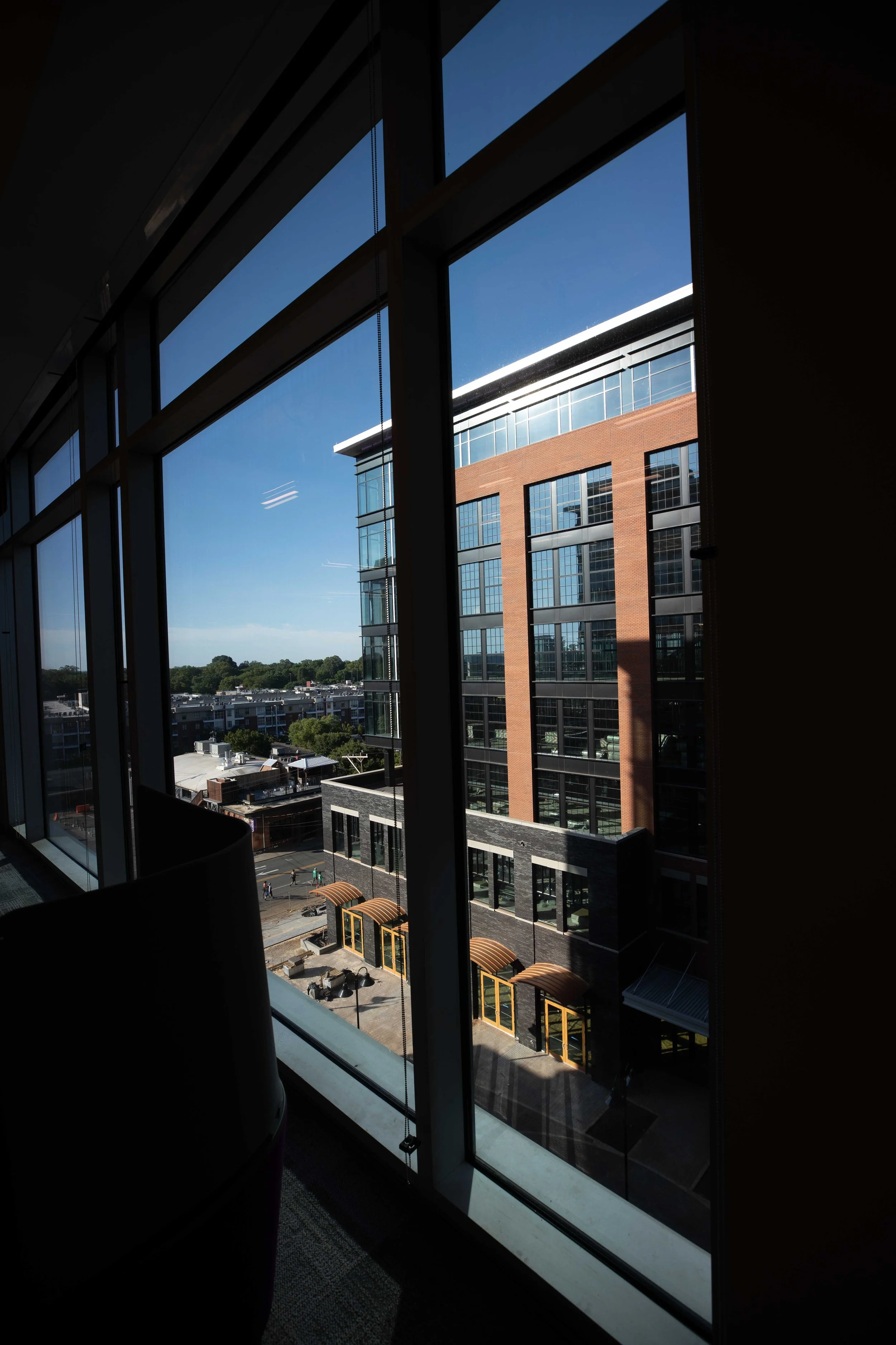 View from a large window in a modern office building, showing a cityscape with multi-story buildings, clear blue sky, and trees in the distance.