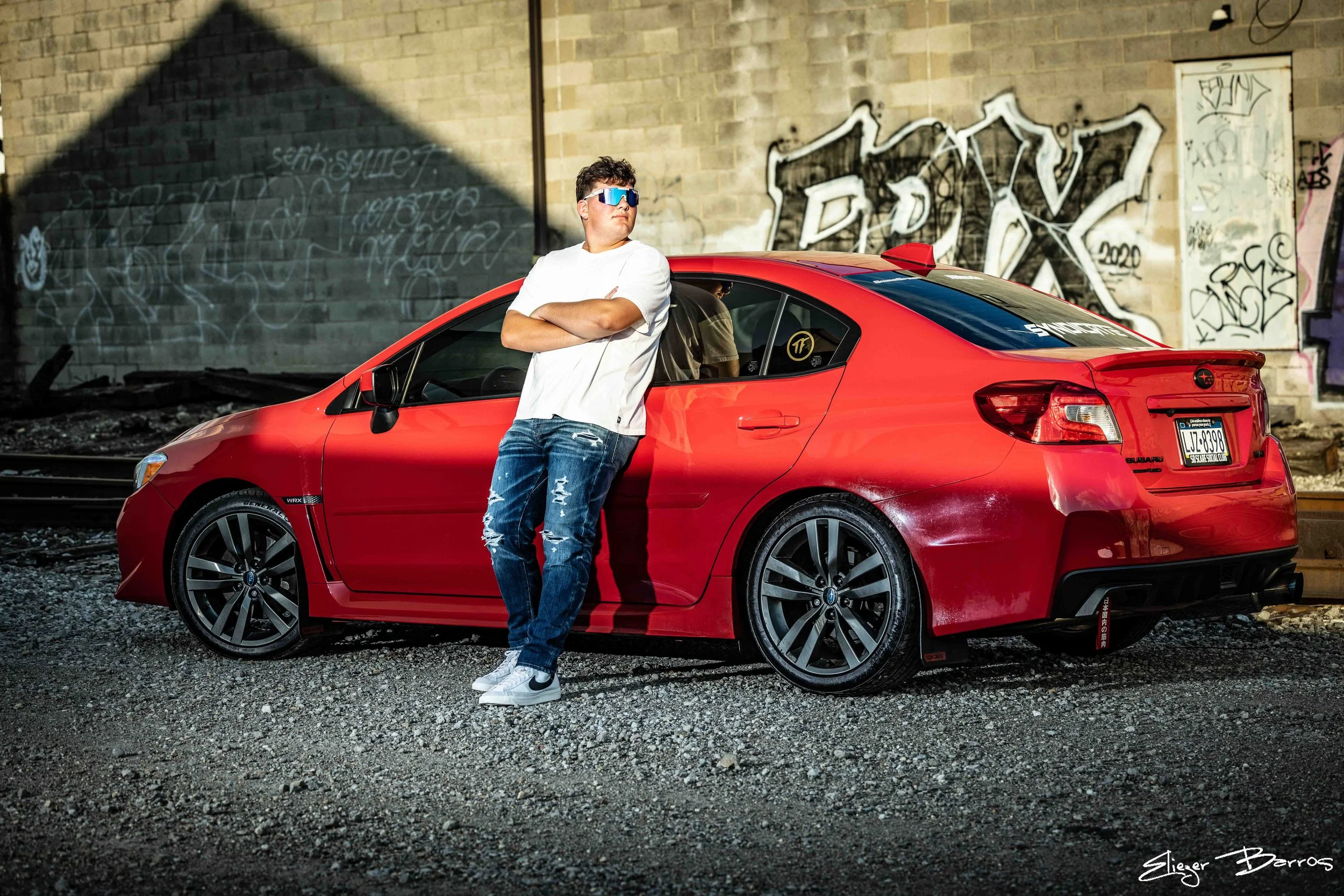 A young man with curly hair wearing sunglasses, a white t-shirt, and ripped jeans stands with crossed arms leaning against a red sports car in front of a graffiti-covered brick wall.