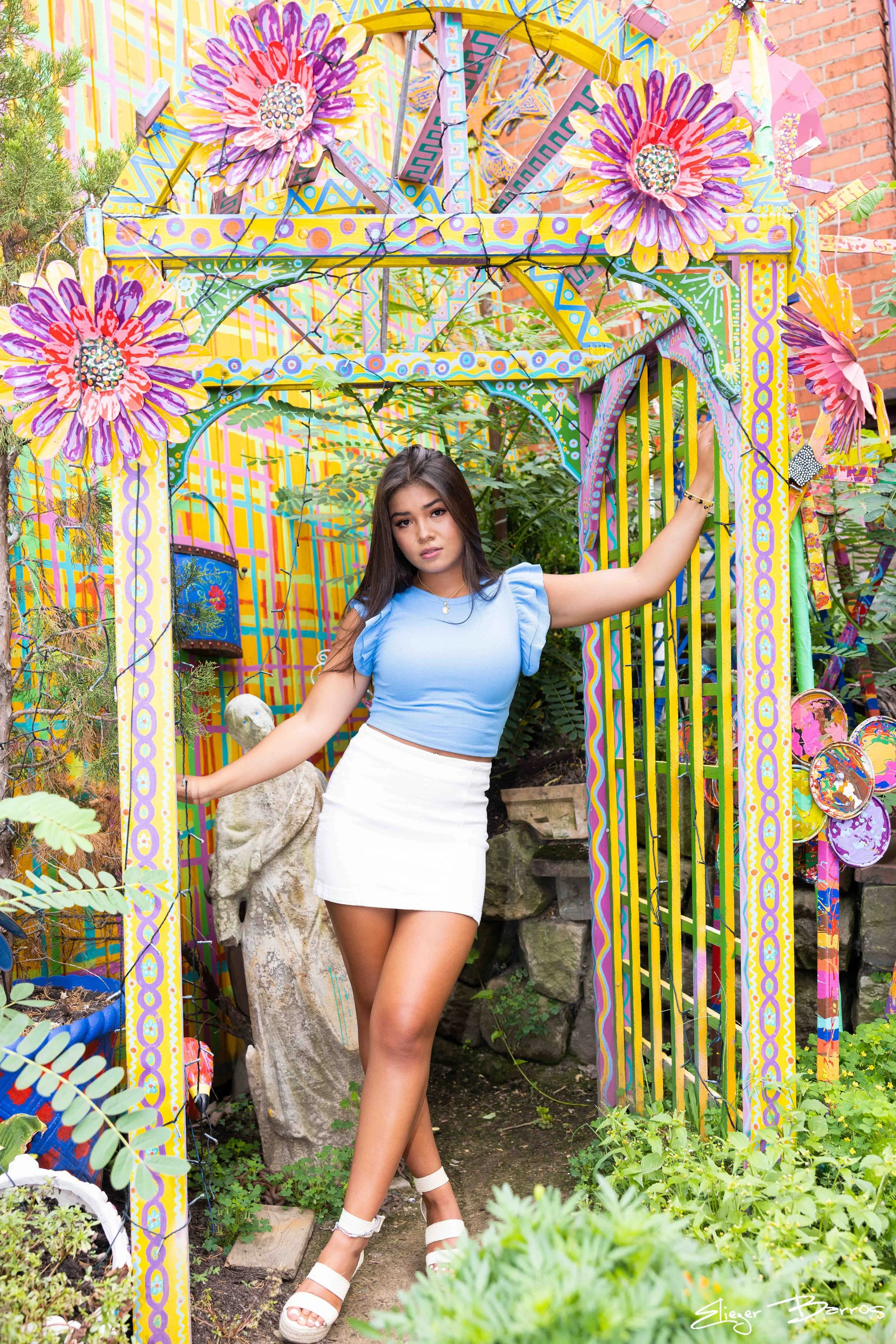 Young woman posing in a colorful, artistic garden setting with painted flowers and decorative elements around her, dressed in a light blue top and white skirt.