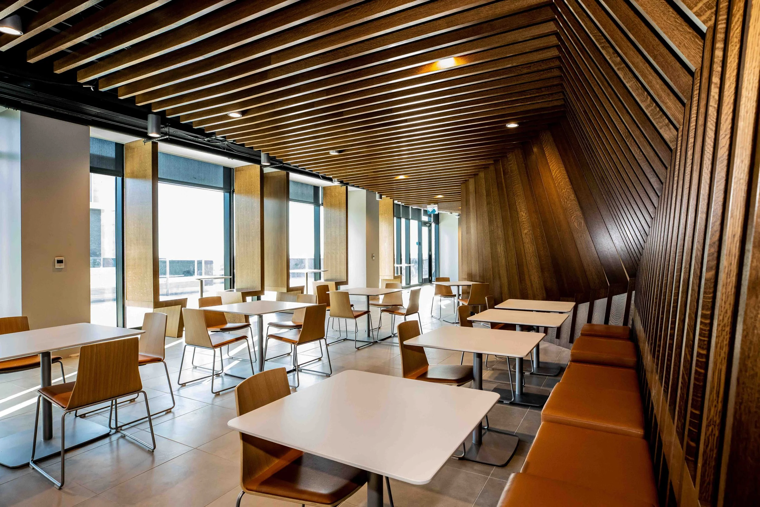 Interior of a modern restaurant or cafe with white tables and wooden chairs, large windows, and a slanted wooden ceiling.