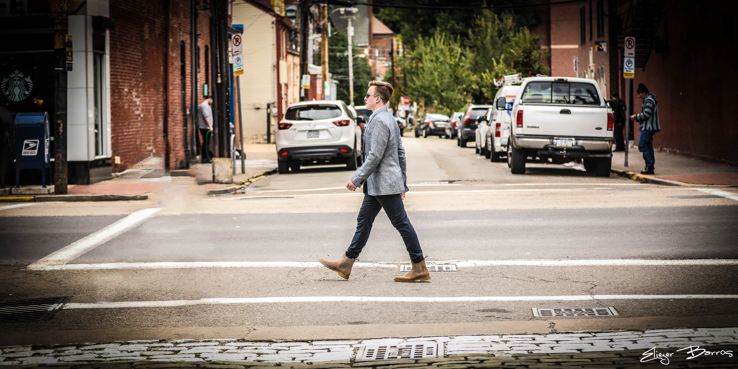 A man in a grey blazer, dark pants, and tan boots walking across a crosswalk on a city street. Behind him, parked cars, buildings, and a person wearing a striped hoodie looking at their phone. The scene shows urban life with trees and a cloudy sky.