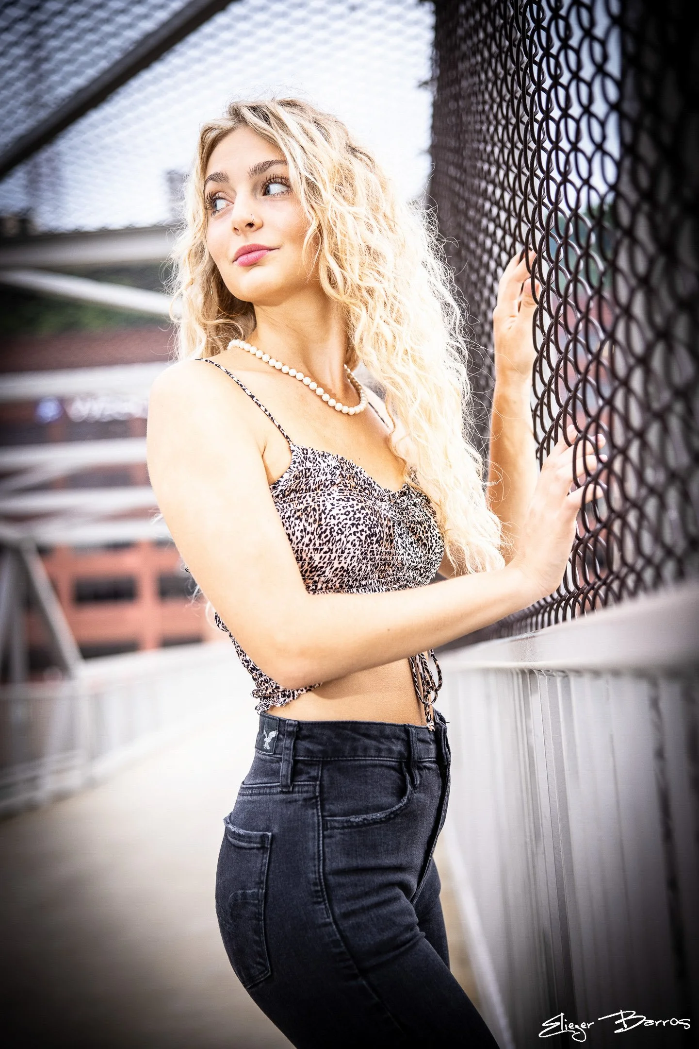 A young woman with long wavy blonde hair is standing near a chain-link fence, holding onto it with her right hand. She is wearing a leopard print crop top and dark high-waisted jeans. She is looking to her left with a slight smile.