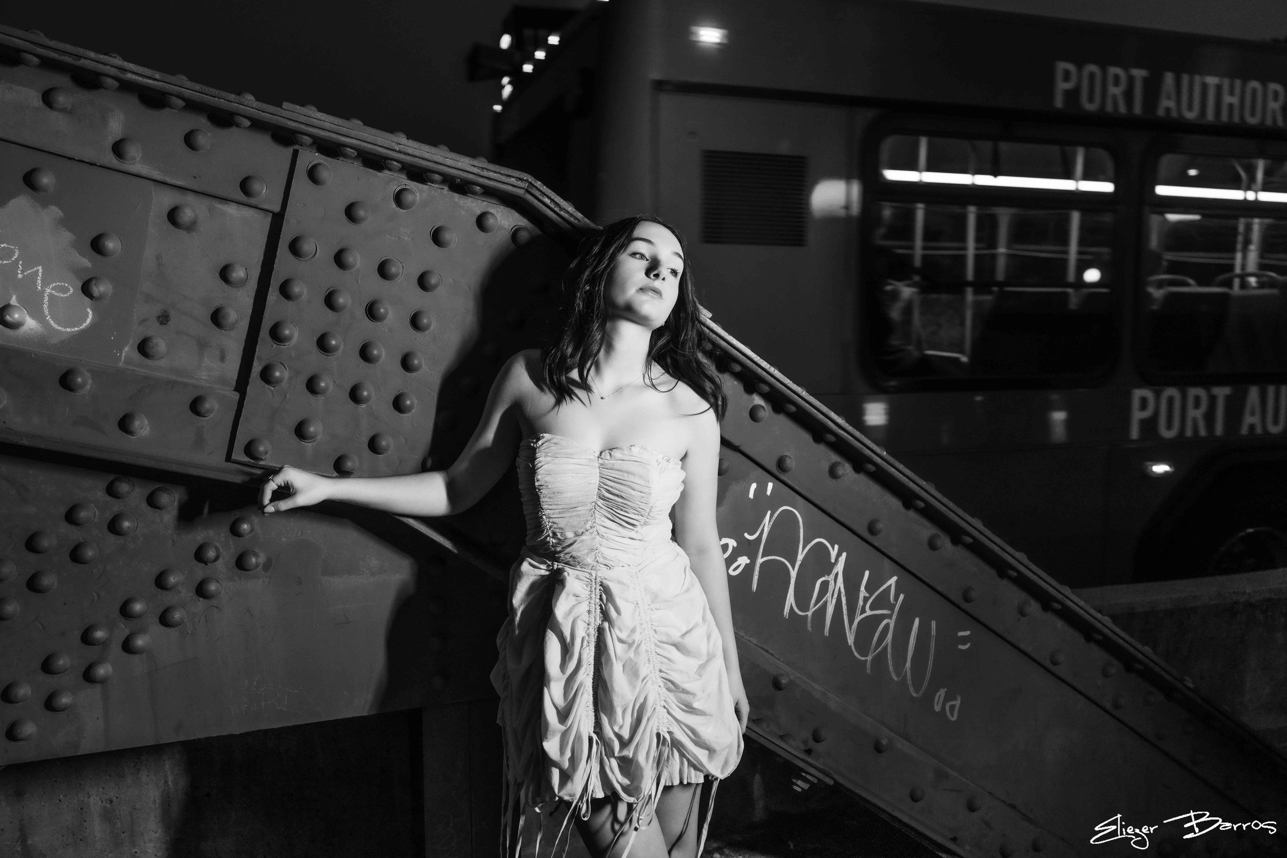 A woman in a strapless dress standing against a textured metal wall at night. The scene is black and white.