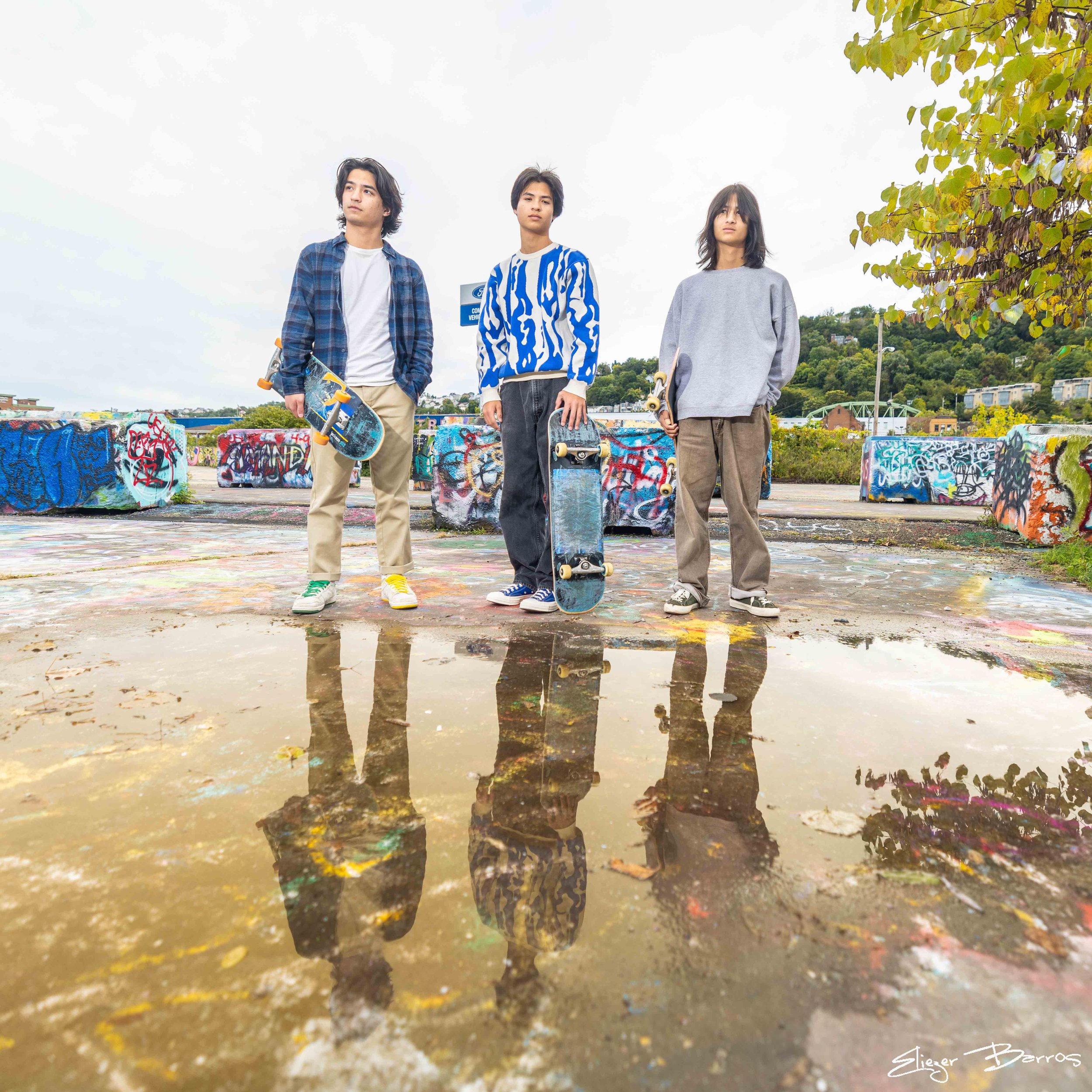 Three young men standing on a graffiti-covered skate park with skateboards under their arms, reflected in a large puddle, with a tree and houses in the background.