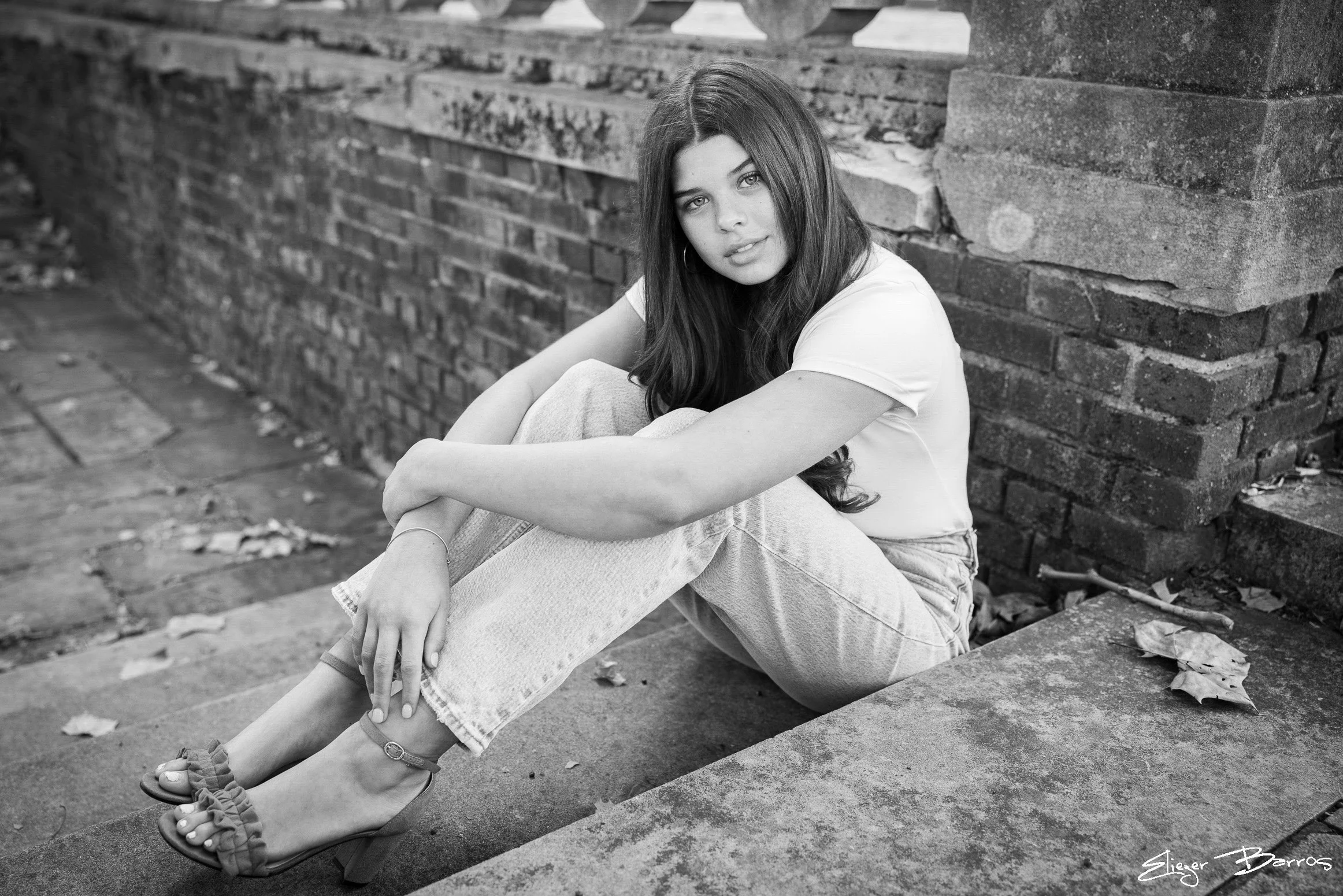 A young woman with long dark hair sitting on a sidewalk next to a brick wall, wearing a white t-shirt and light-colored jeans, gazing at the camera.