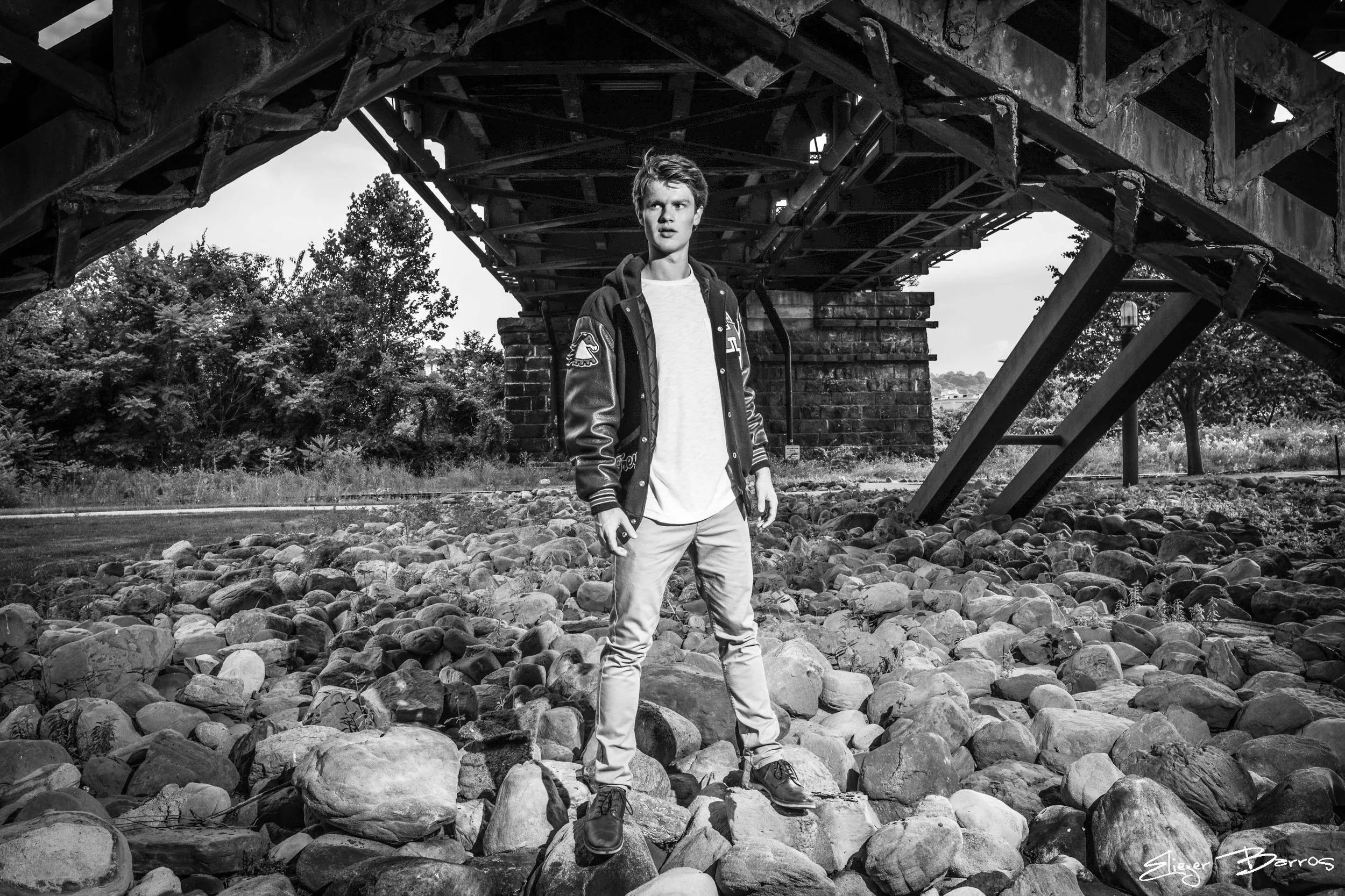 A young man stands on a rocky ground beneath a large, industrial bridge structure, looking off into the distance with a serious expression. The image is in black and white.