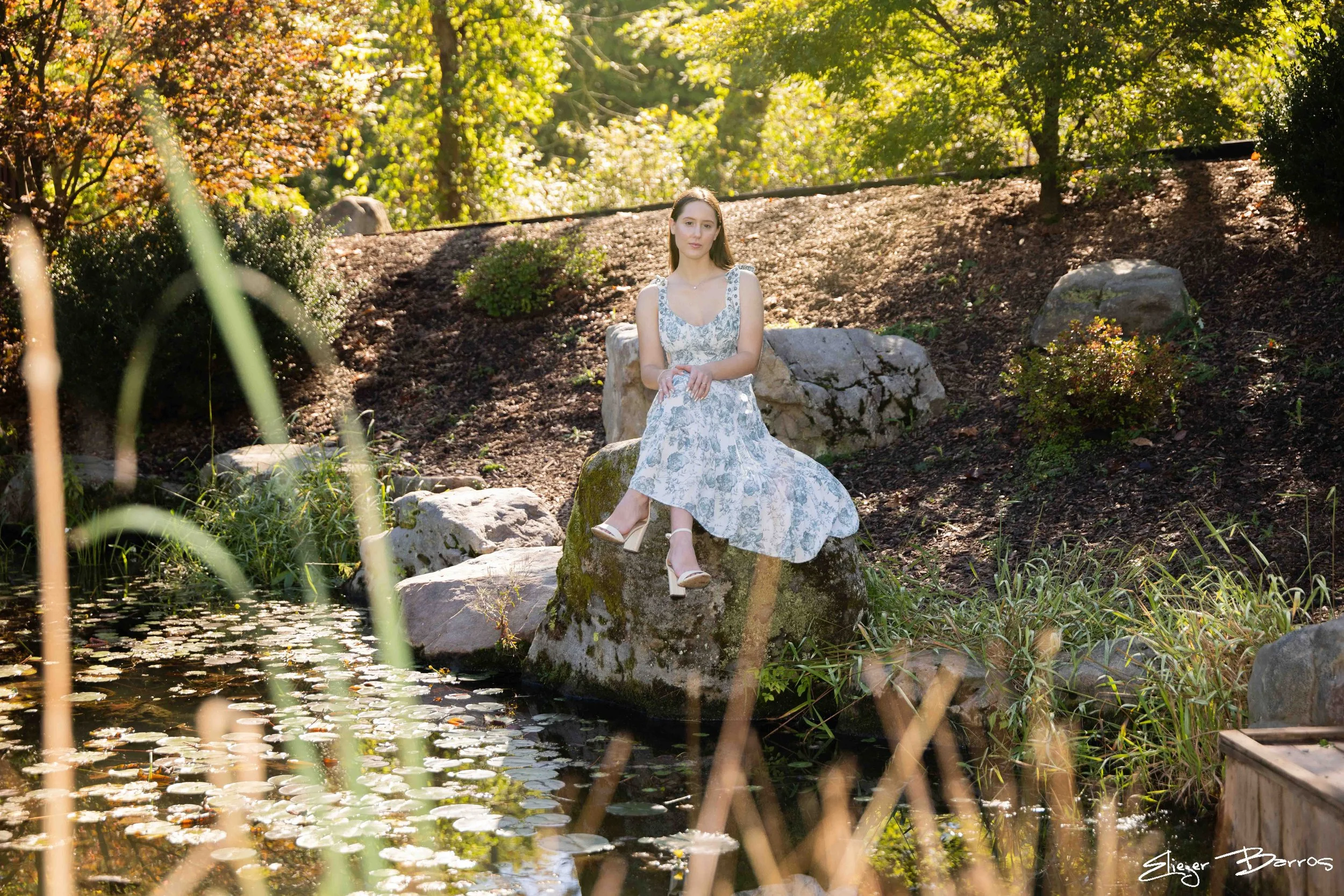 A young woman in a white patterned dress and high heels sitting on a large rock by a pond in a wooded area during daylight, surrounded by greenery and trees.