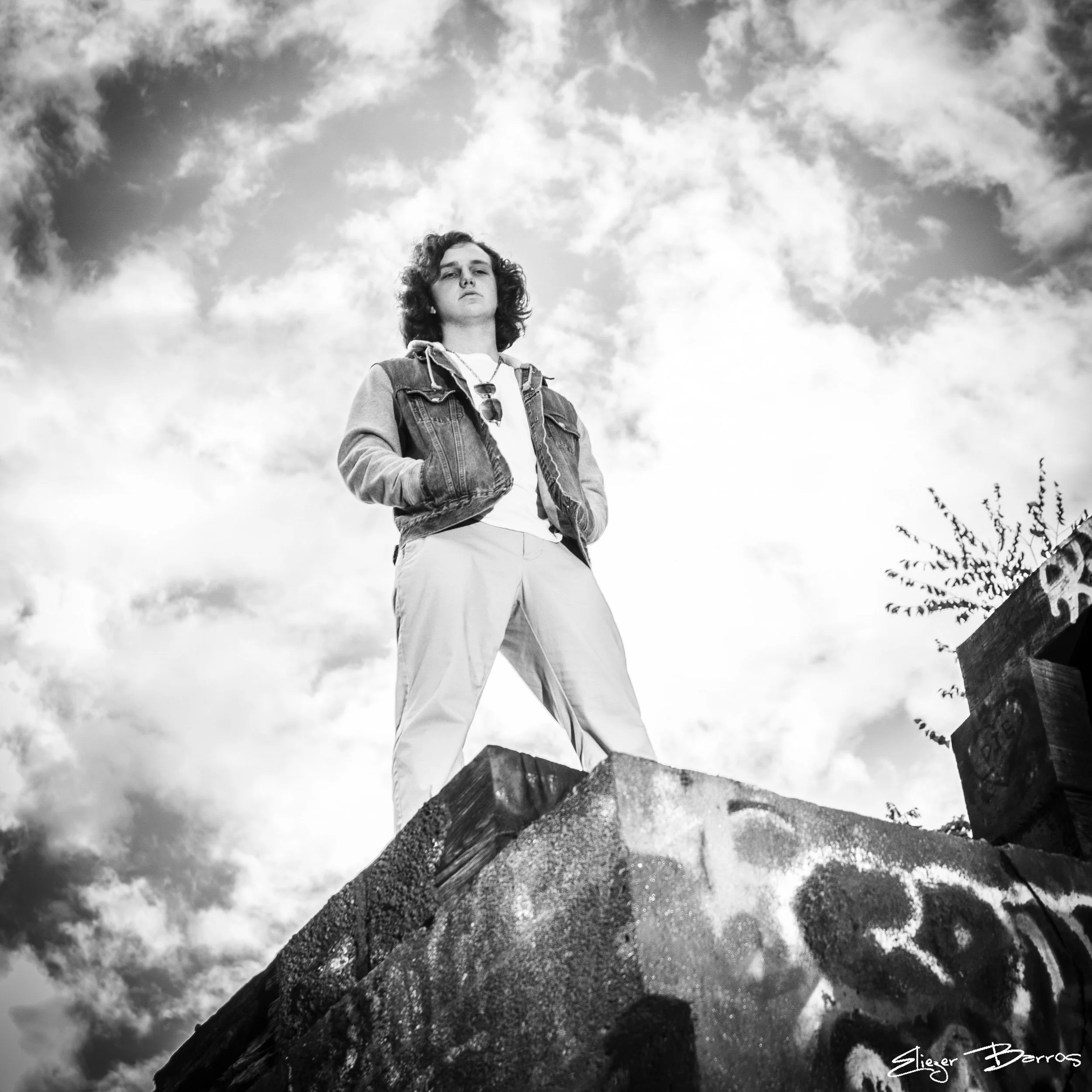 Black-and-white photo of a young man with curly hair standing outdoors against a sky with scattered clouds, wearing a denim jacket and a white shirt, with mountains and trees in the background.