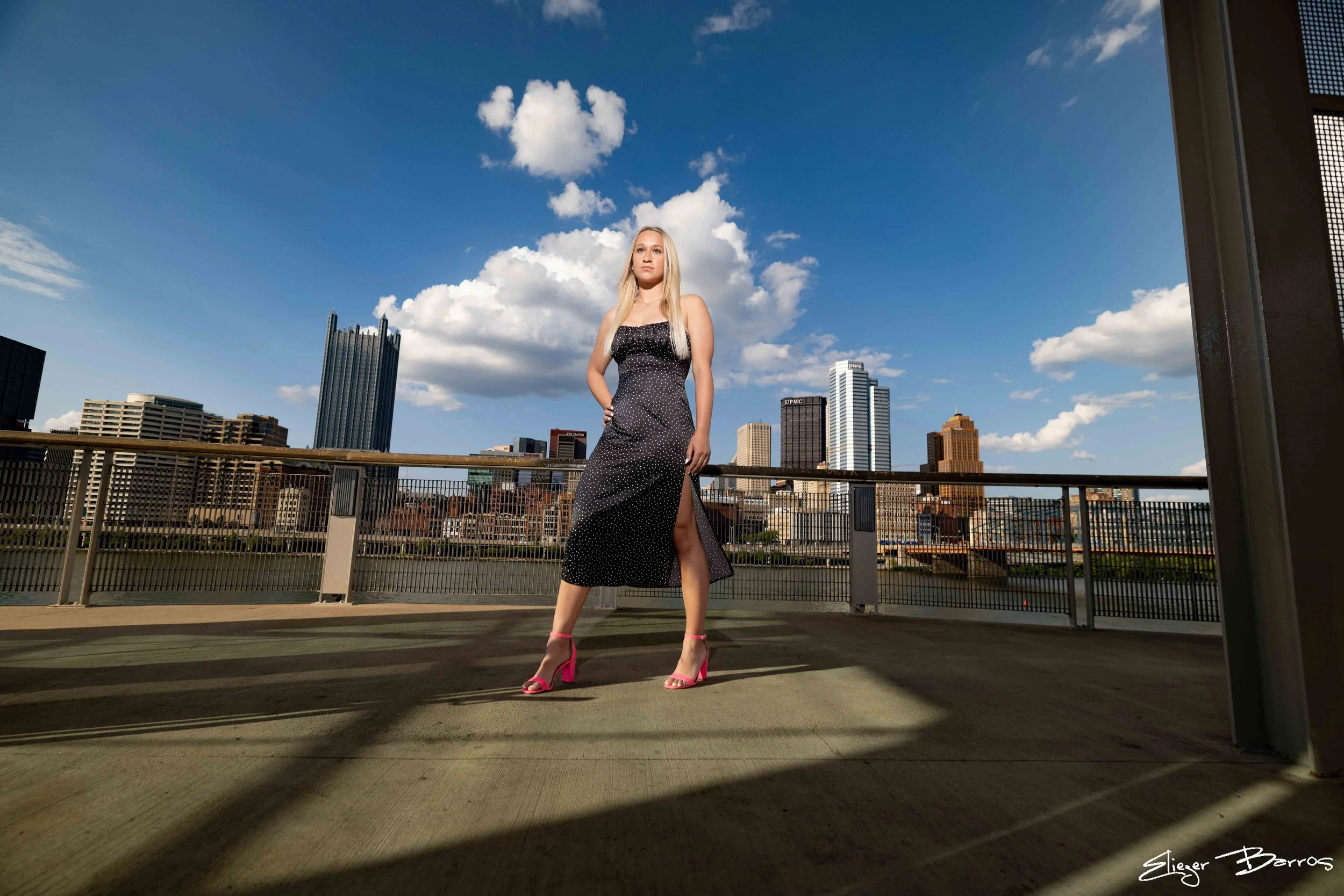 A young woman in a black dress with white polka dots, high heels, and blonde hair stands on a rooftop parking lot against a city skyline with tall buildings and a partly cloudy sky.