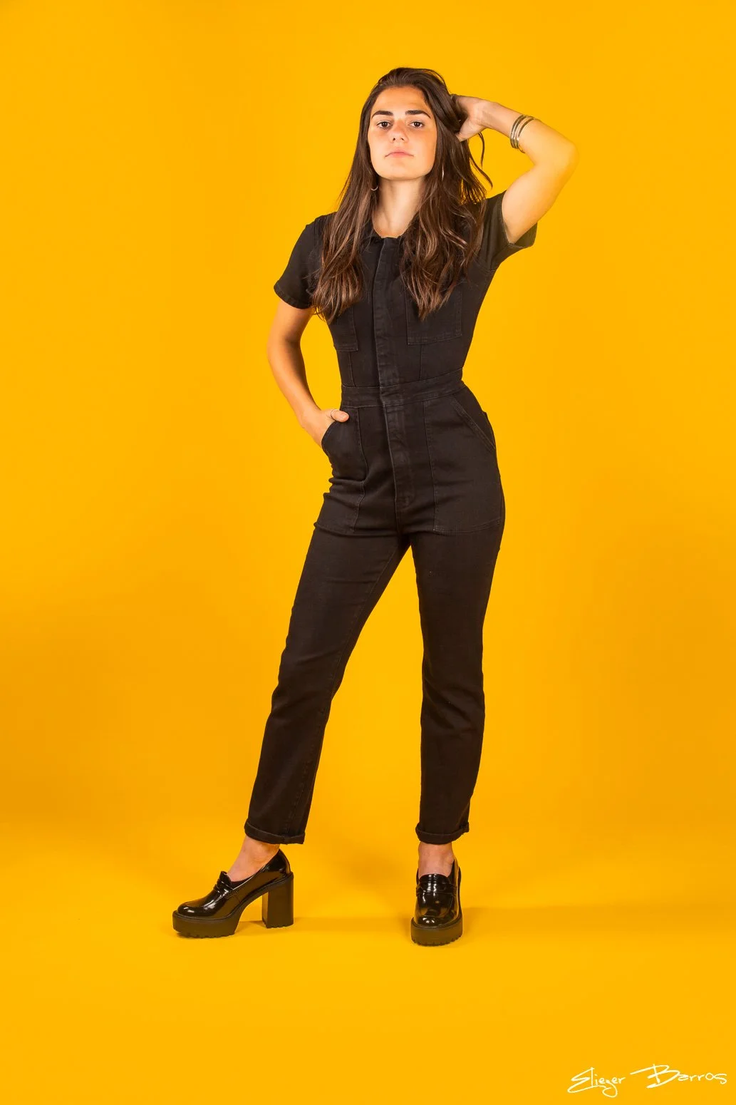 A woman in a black jumpsuit and black platform shoes stands against a bright yellow background, with one hand on her hip and the other behind her head.