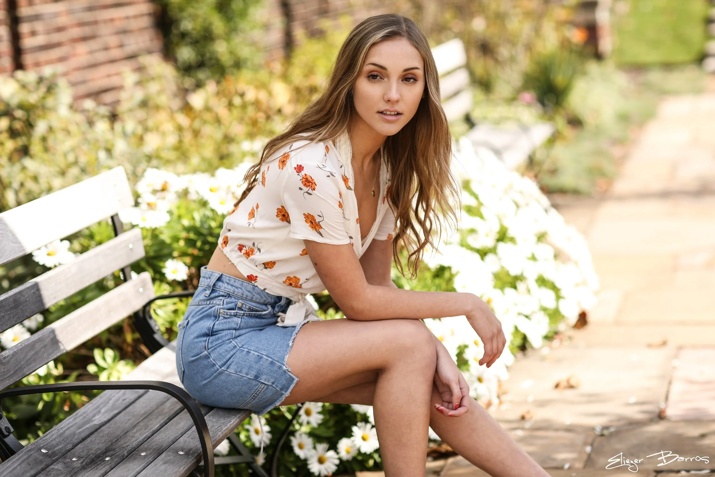 A young woman with long wavy hair sitting on a park bench surrounded by white flowers and green foliage. She is wearing a white floral top and denim shorts, looking slightly to the side with a relaxed expression.