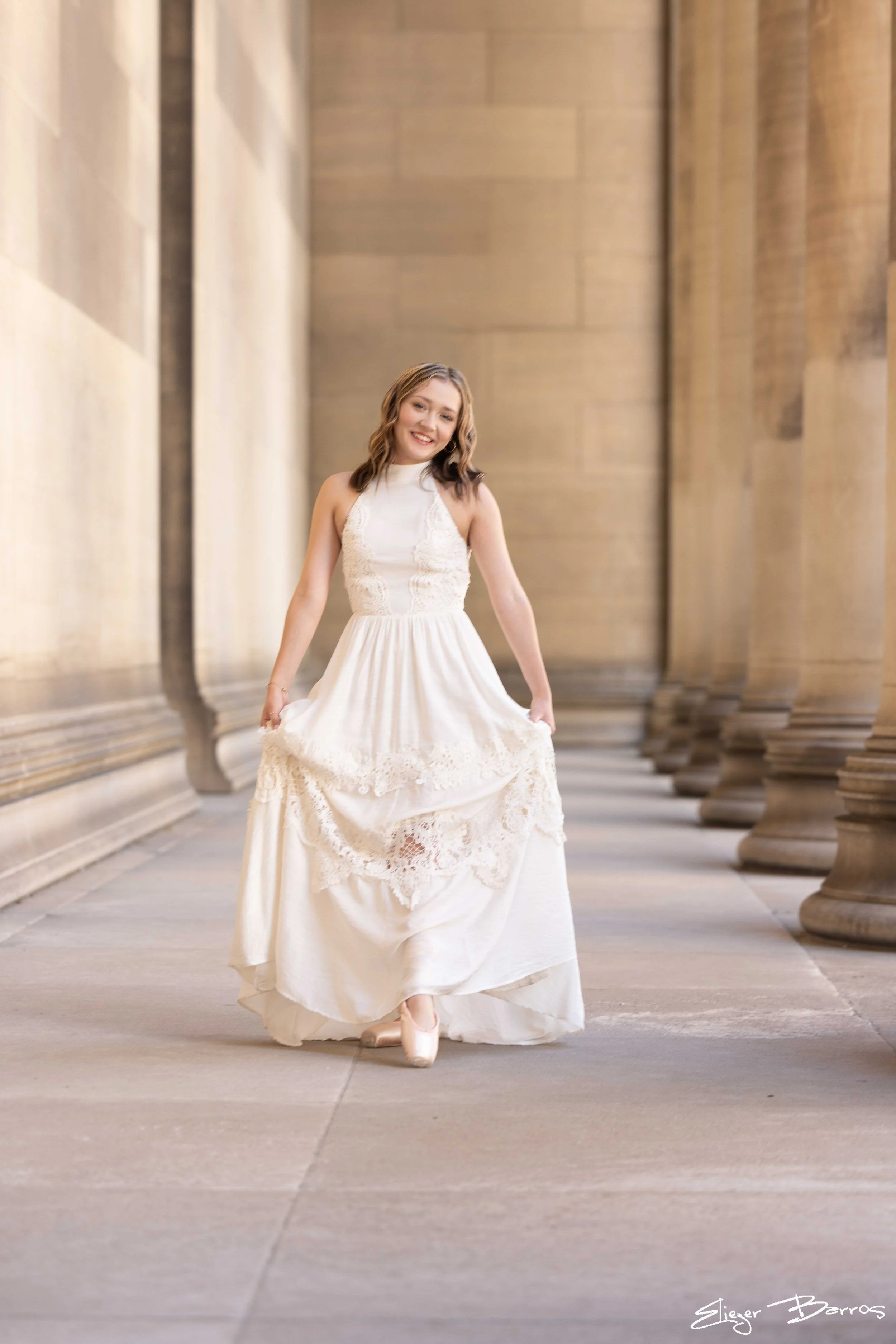 A young woman in a white gown is walking in a corridor lined with large stone columns, smiling and holding her dress as she moves forward.