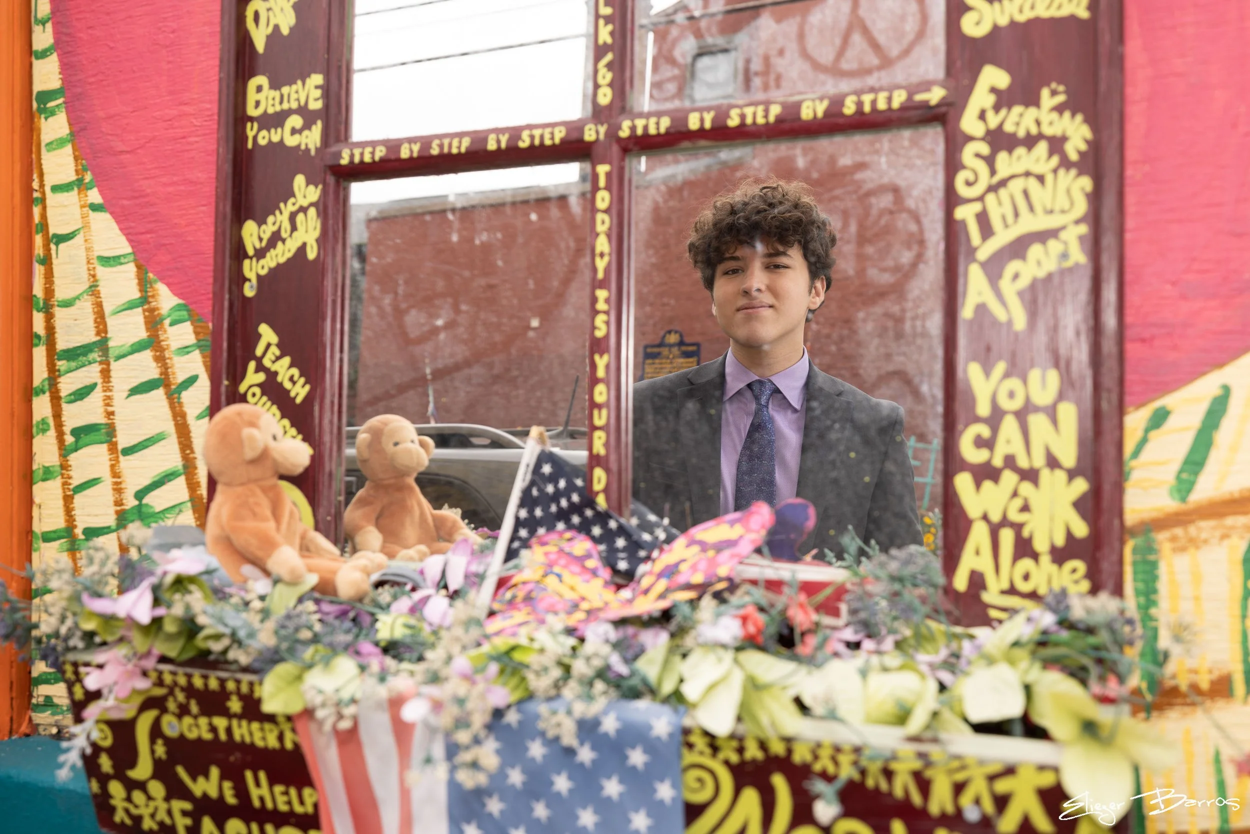 A man with curly hair and a suit looking at a colorful display of toys, stuffed animals, and flowers inside a decorated frame with yellow motivational words and phrases written on it.