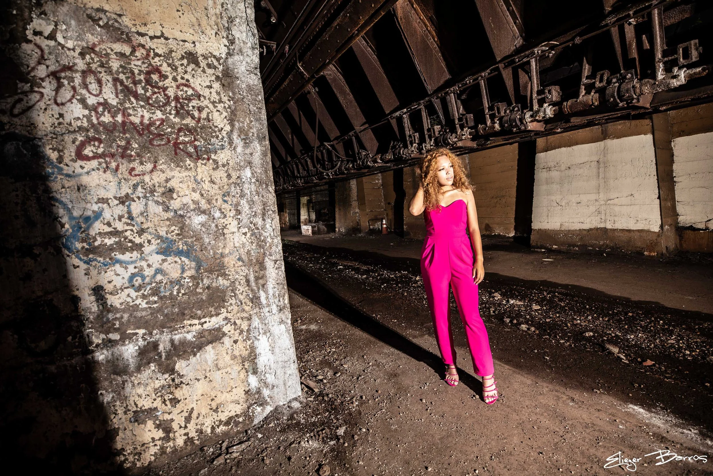 A woman with curly hair standing in an abandoned industrial space, wearing a bright pink jumpsuit and high-heeled sandals, with graffiti on a concrete pillar nearby.