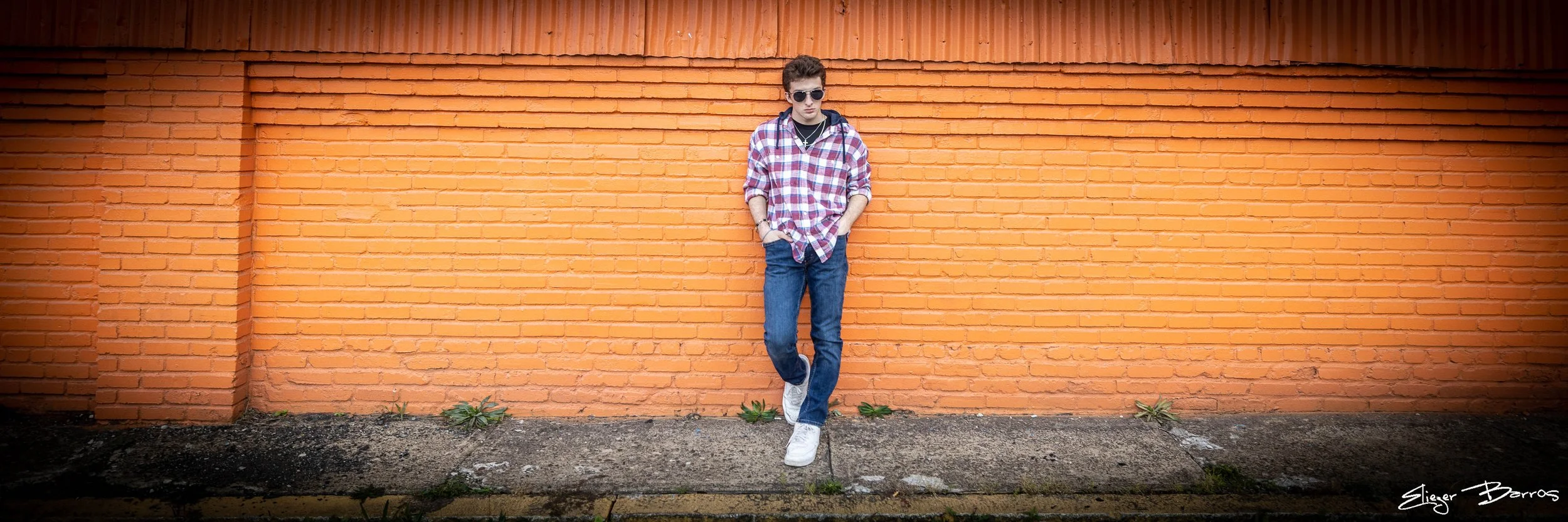 A young man with sunglasses, wearing a plaid shirt, jeans, and white sneakers, leaning against an orange brick wall outdoors.