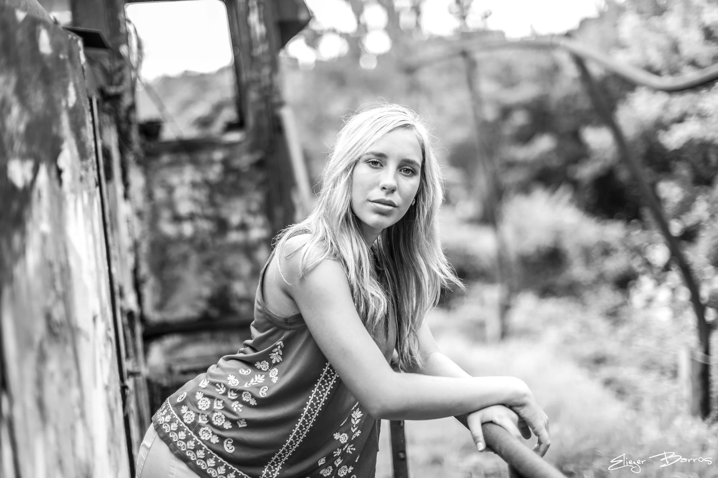A young woman with long blonde hair sitting outdoors near a wooden structure, wearing a patterned sleeveless top, with a blurred natural background in black and white.