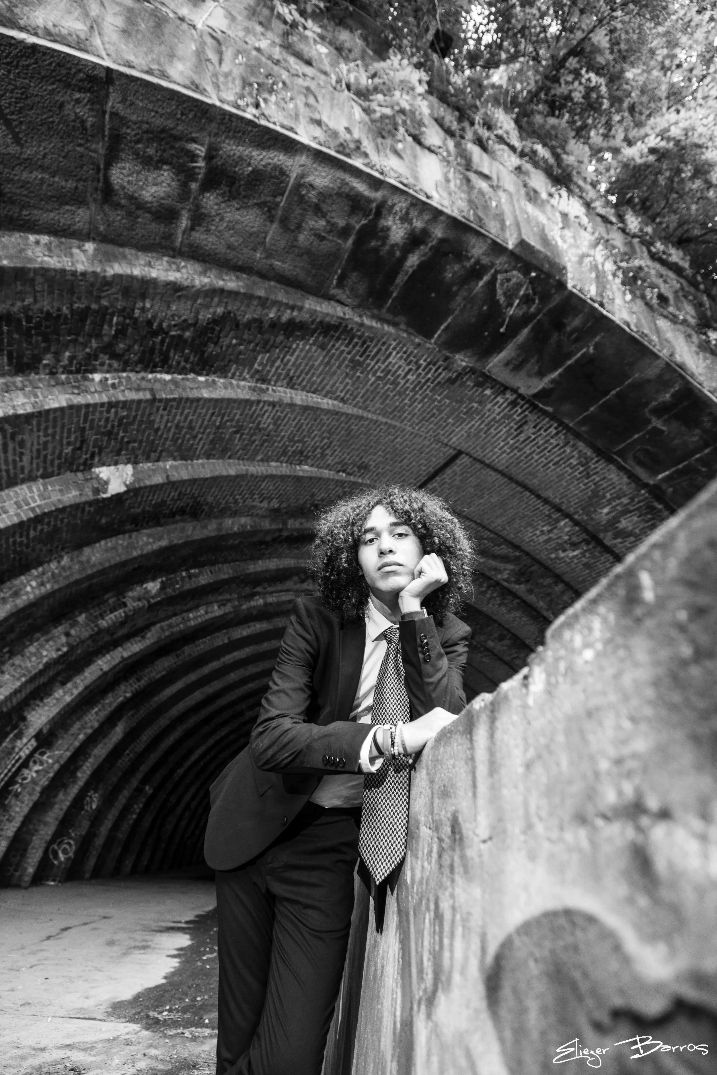 A woman in a suit and tie leaning on a concrete wall under a tunnel with brick and stone archways, looking at the camera.
