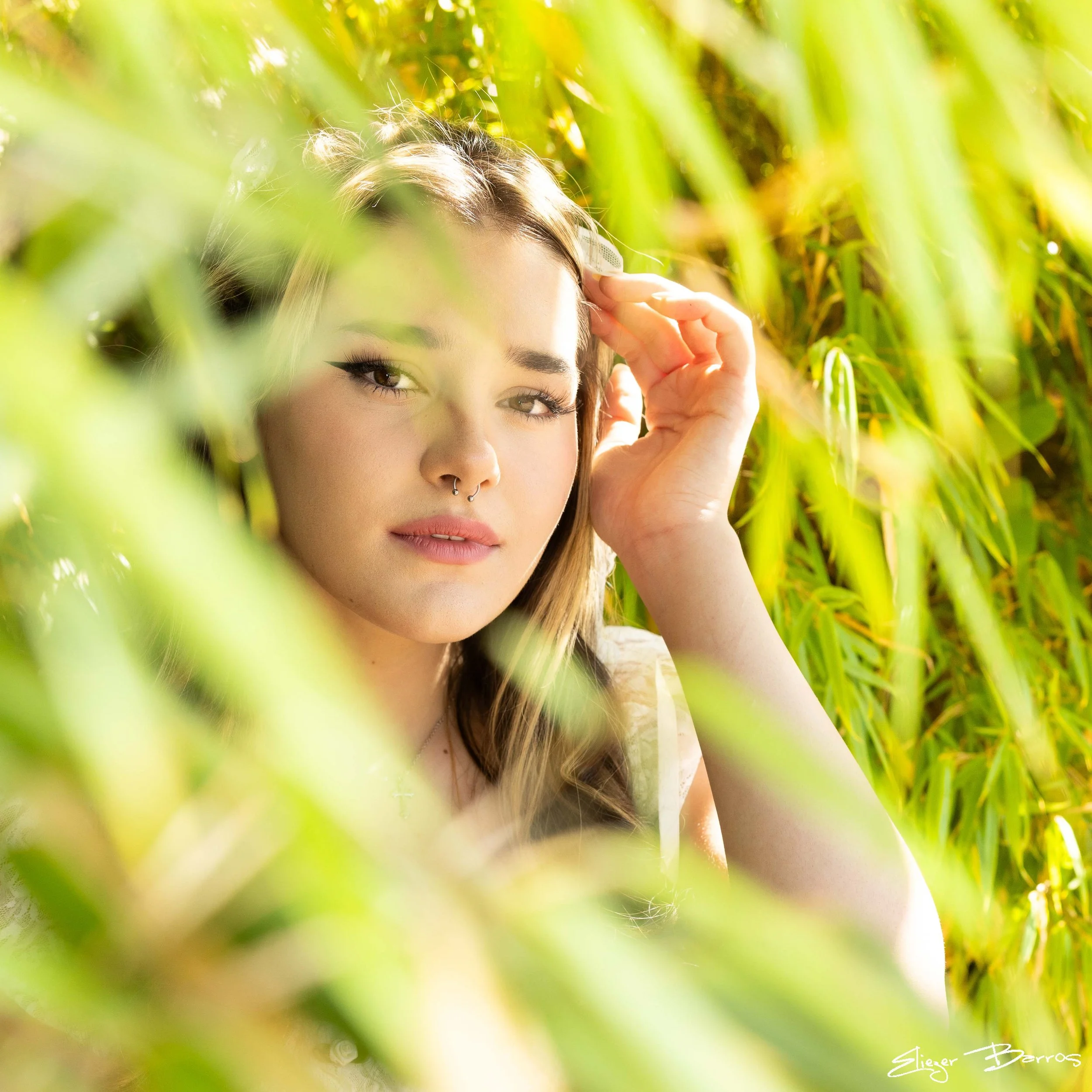 A woman with brown hair, eye makeup, and piercings around her nose is partially visible through tall green grass, with sunlight highlighting her face, in an outdoor setting.