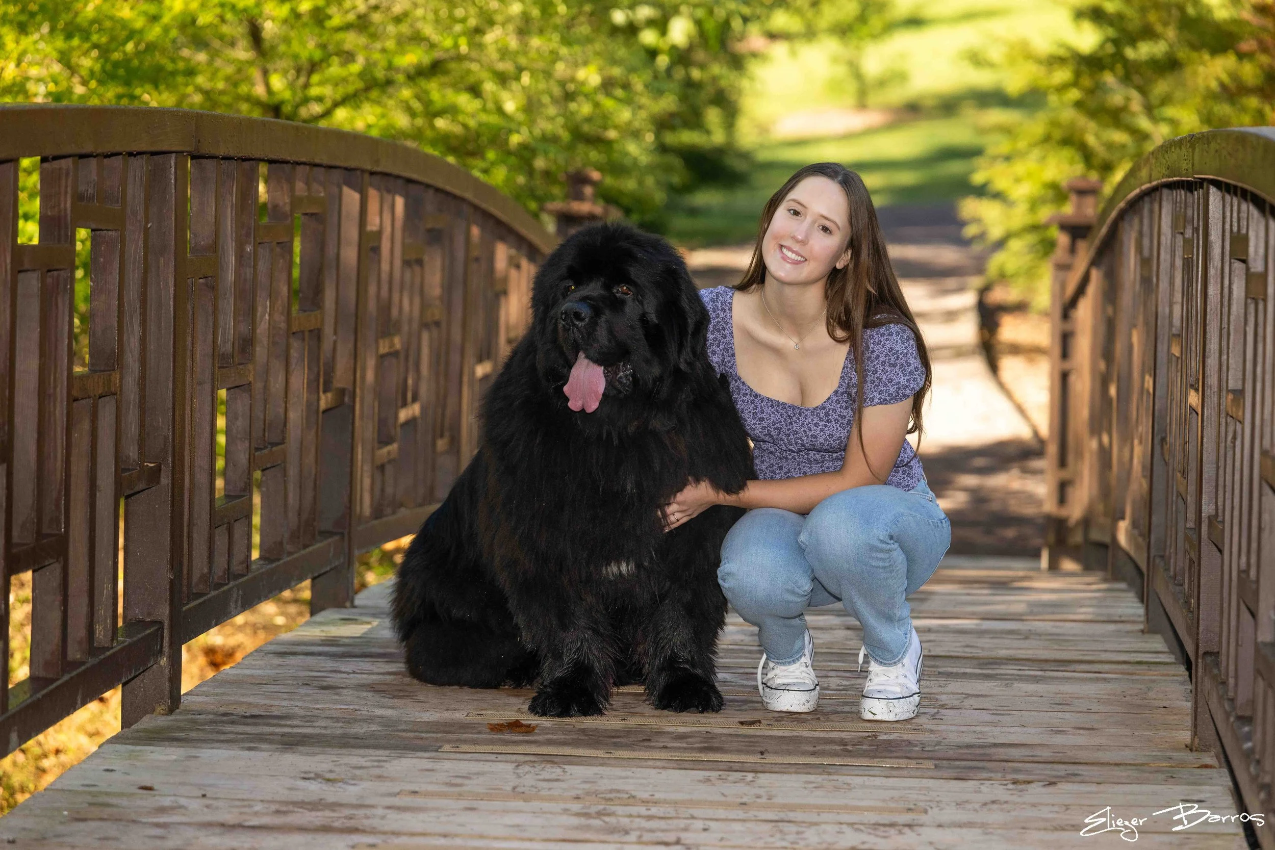 A young woman squatting on a wooden bridge with a large black Newfoundland dog sitting beside her, both smiling amid green trees.