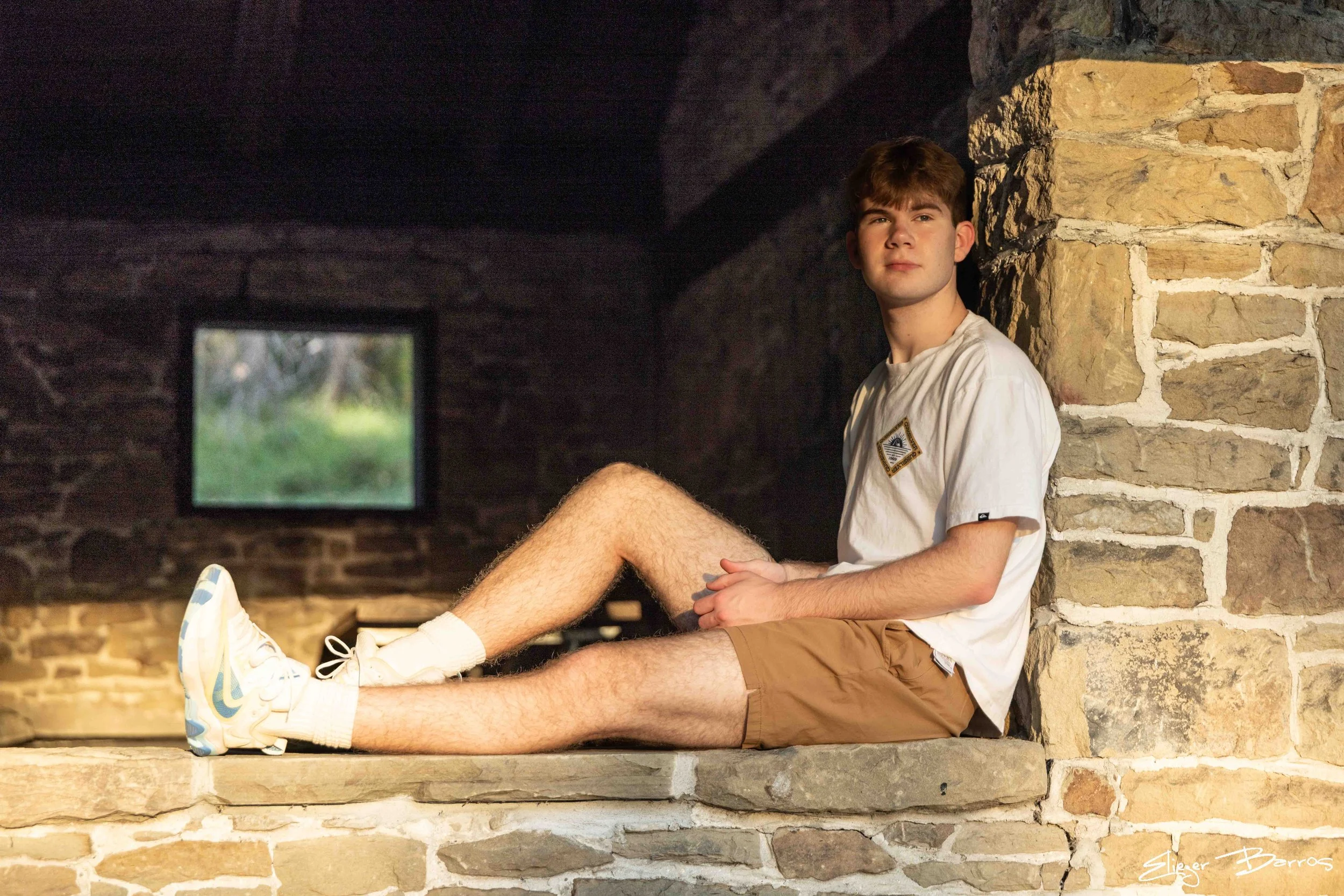 A young man with brown hair wearing a white t-shirt and khaki shorts sitting on a brick ledge against a brick wall, with a window in the background showing greenery outside.