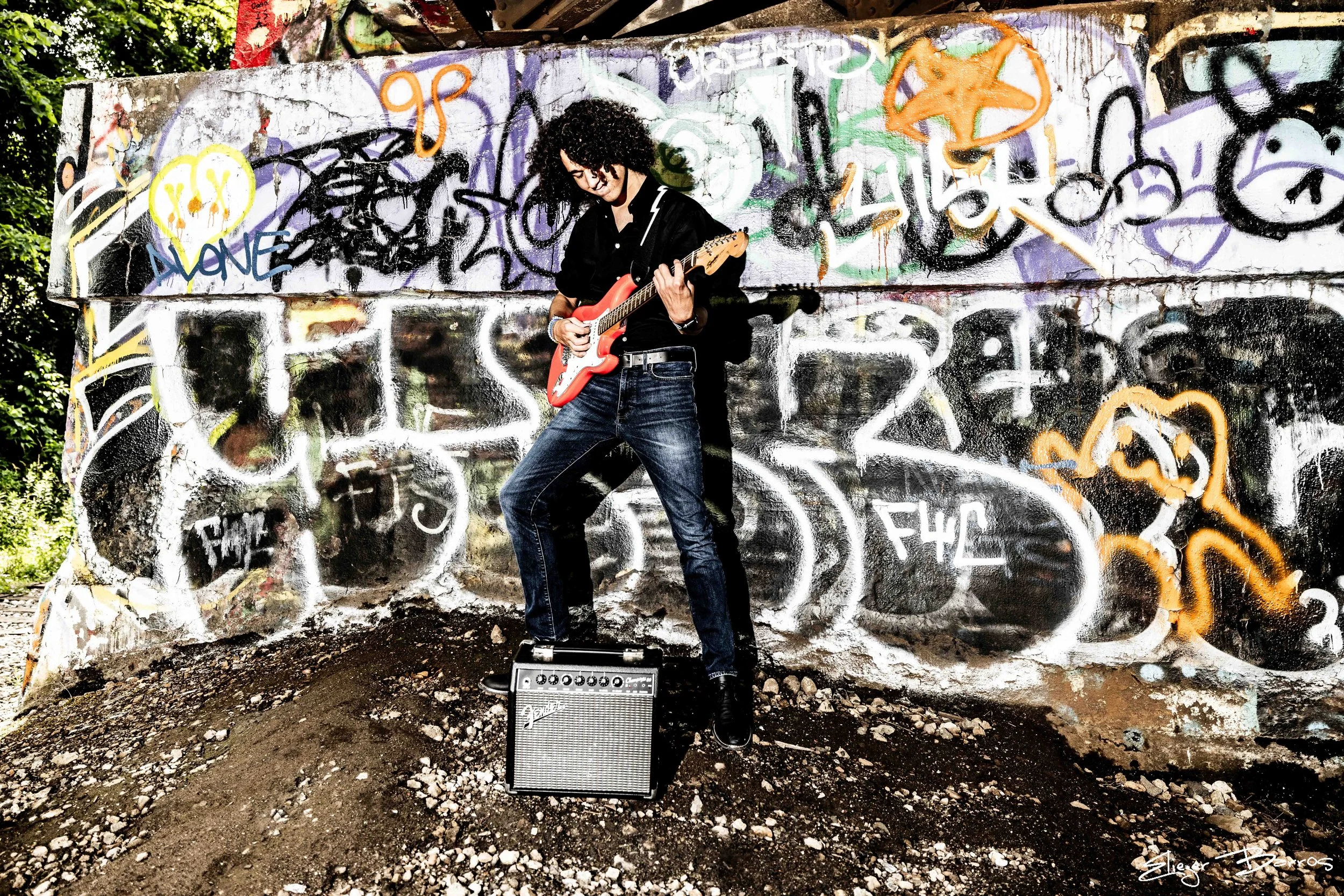 A person with curly hair playing an electric guitar in front of a graffiti-covered wall, with an amplifier on the ground in front of them.