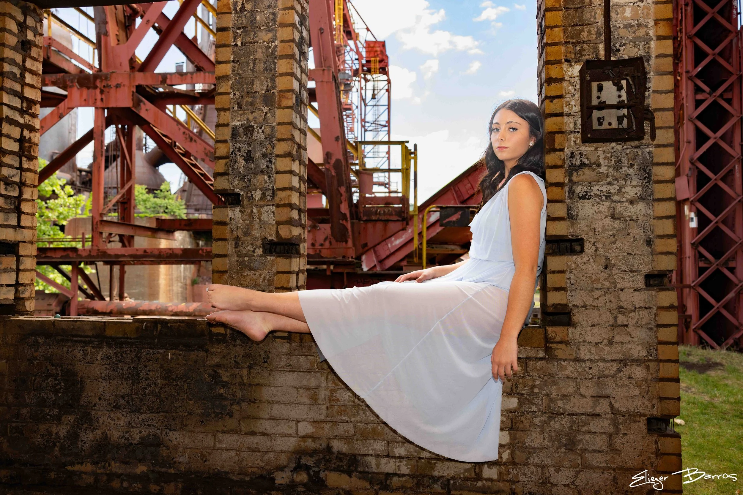 A woman in a white dress sitting on a brick ledge between brick columns with industrial red metal structures in the background on a partly cloudy day.
