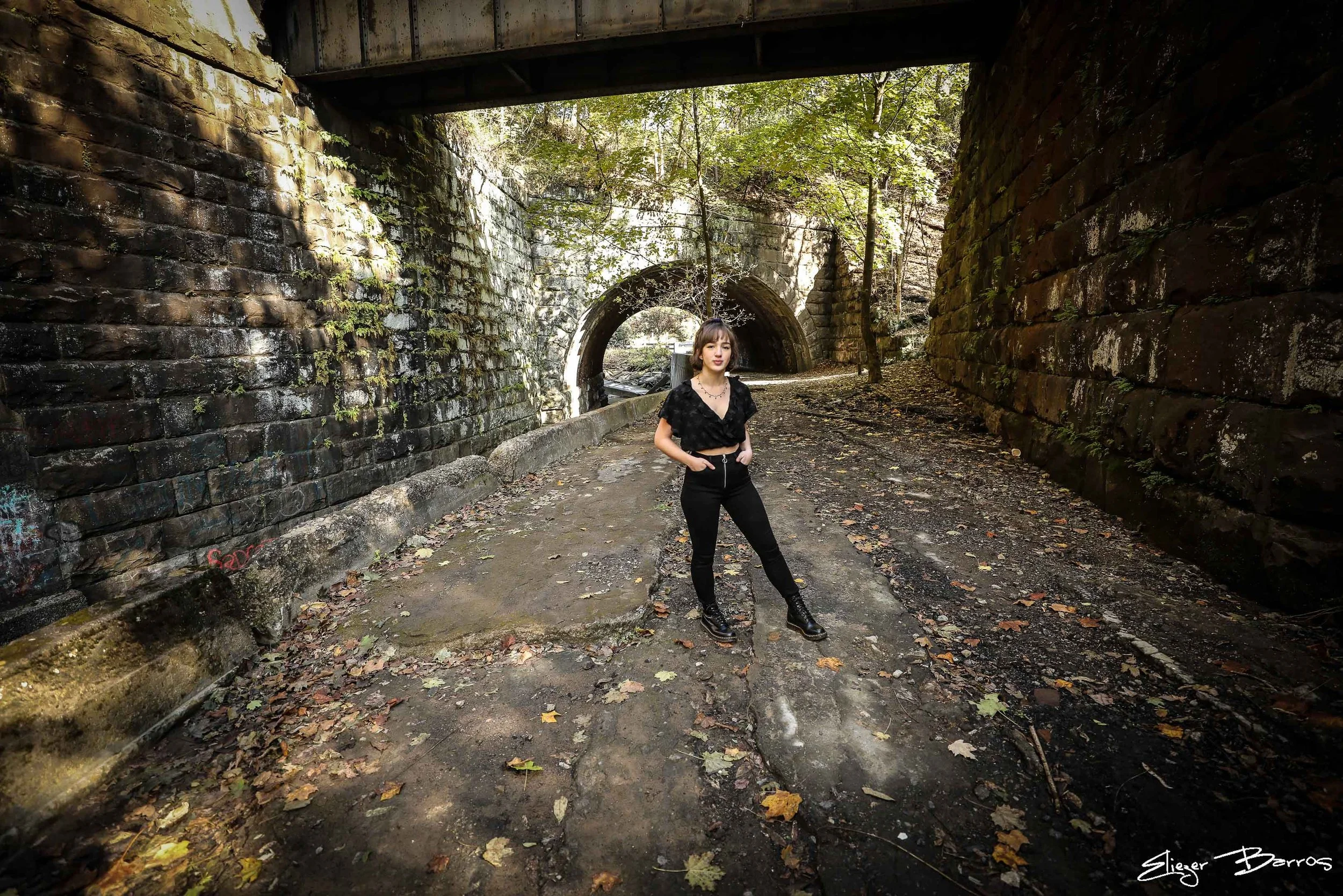 A young woman with short hair wearing a black crop top, black pants, and black combat boots standing on a dirt path beneath a bridge in a wooded area with trees and fallen leaves.