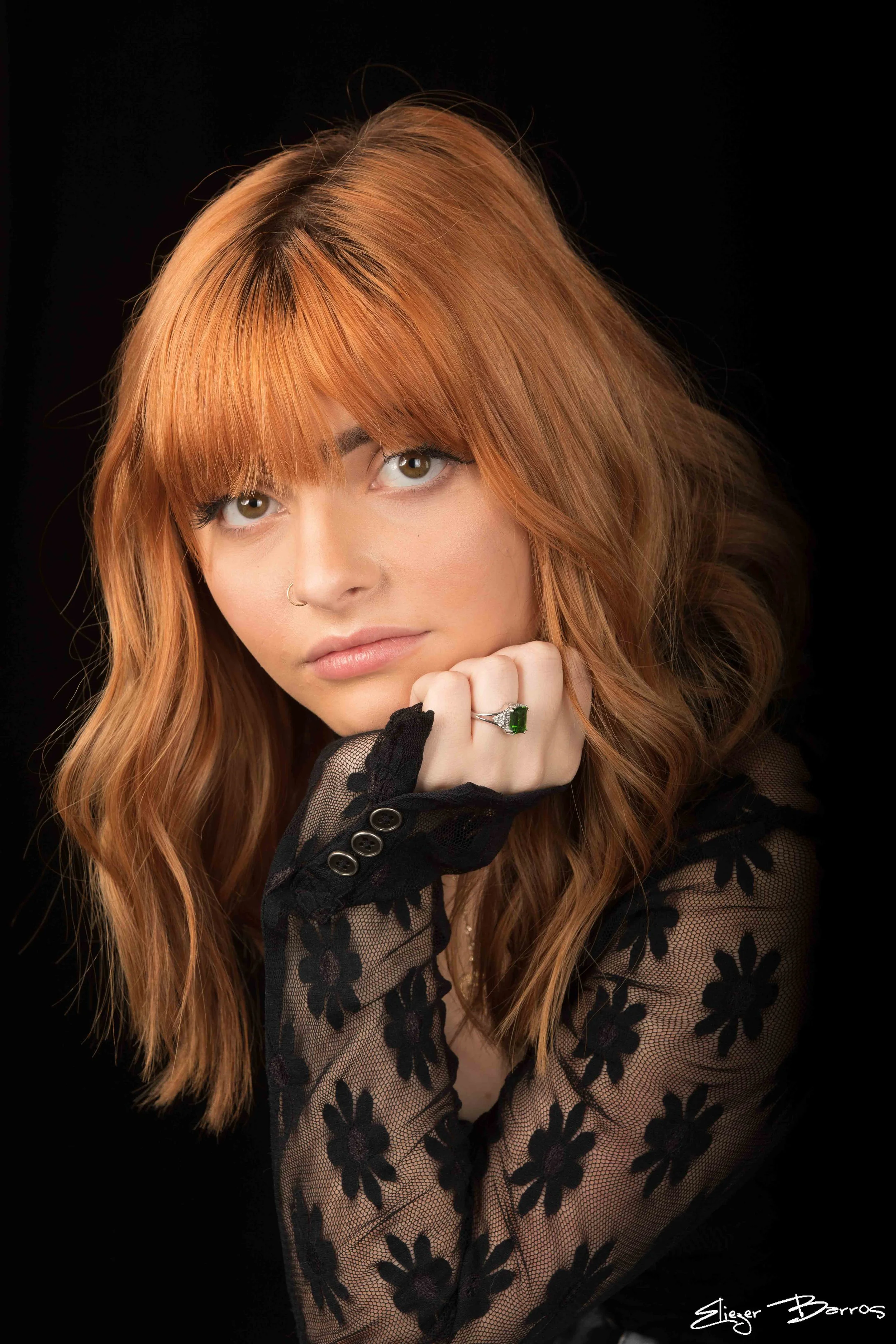 A woman with long, wavy red hair and hazel eyes, wearing a black lace top with floral patterns, a ring with a large green gemstone, and a nose ring, resting her chin on her hand against a black background.