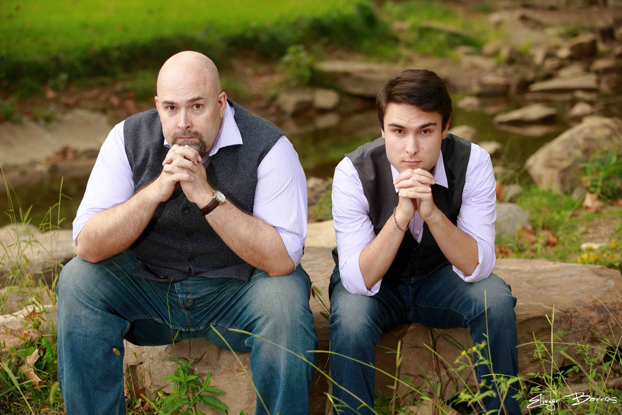 Two men sitting outdoors on a tree trunk by a creek, resting their chins on their hands, looking at the camera.