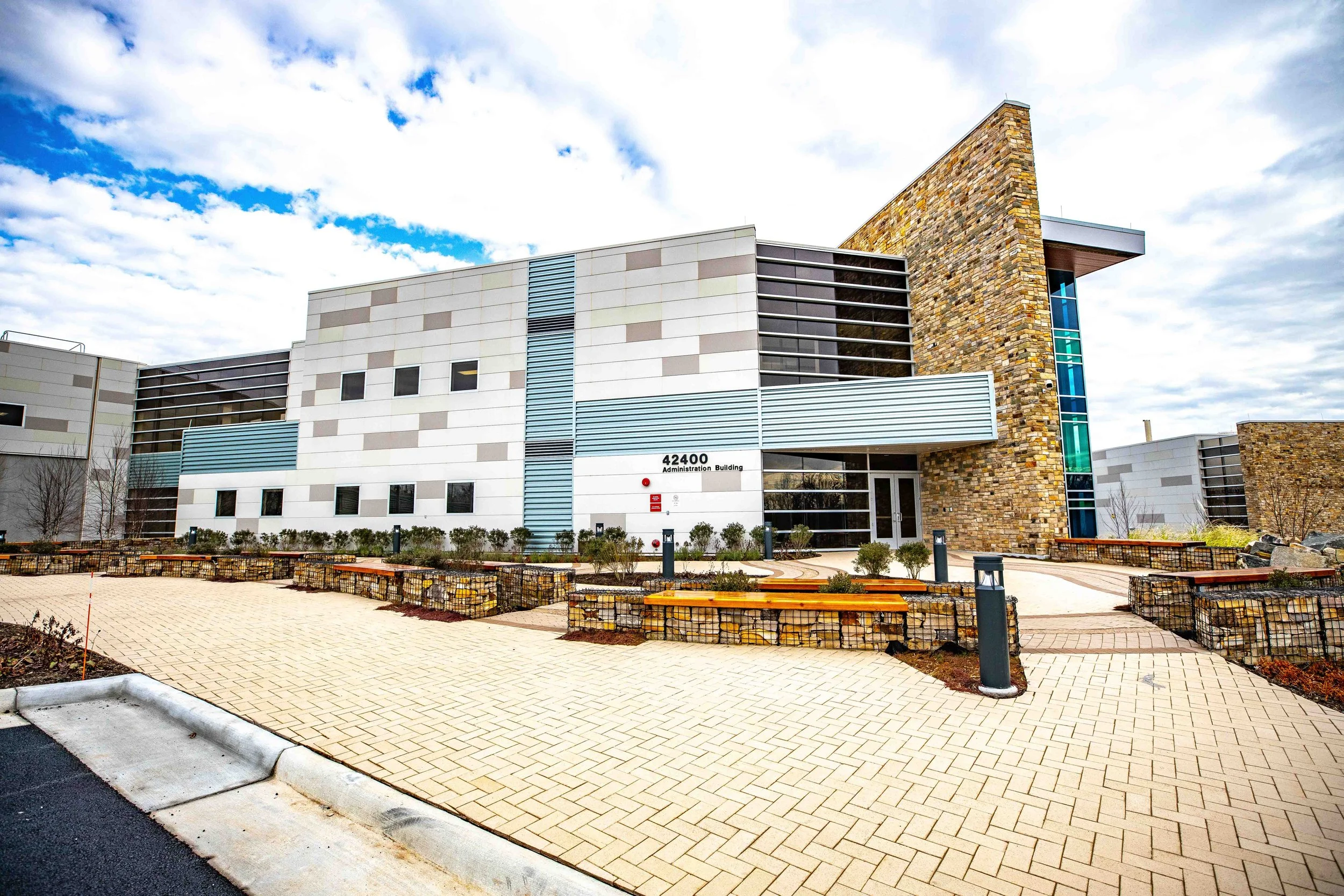 Exterior view of a modern administration building with a stone and metal facade, labeled 42400 Administration Building. The foreground has a paved walkway, benches, and landscaped areas with small bushes under a partly cloudy sky.