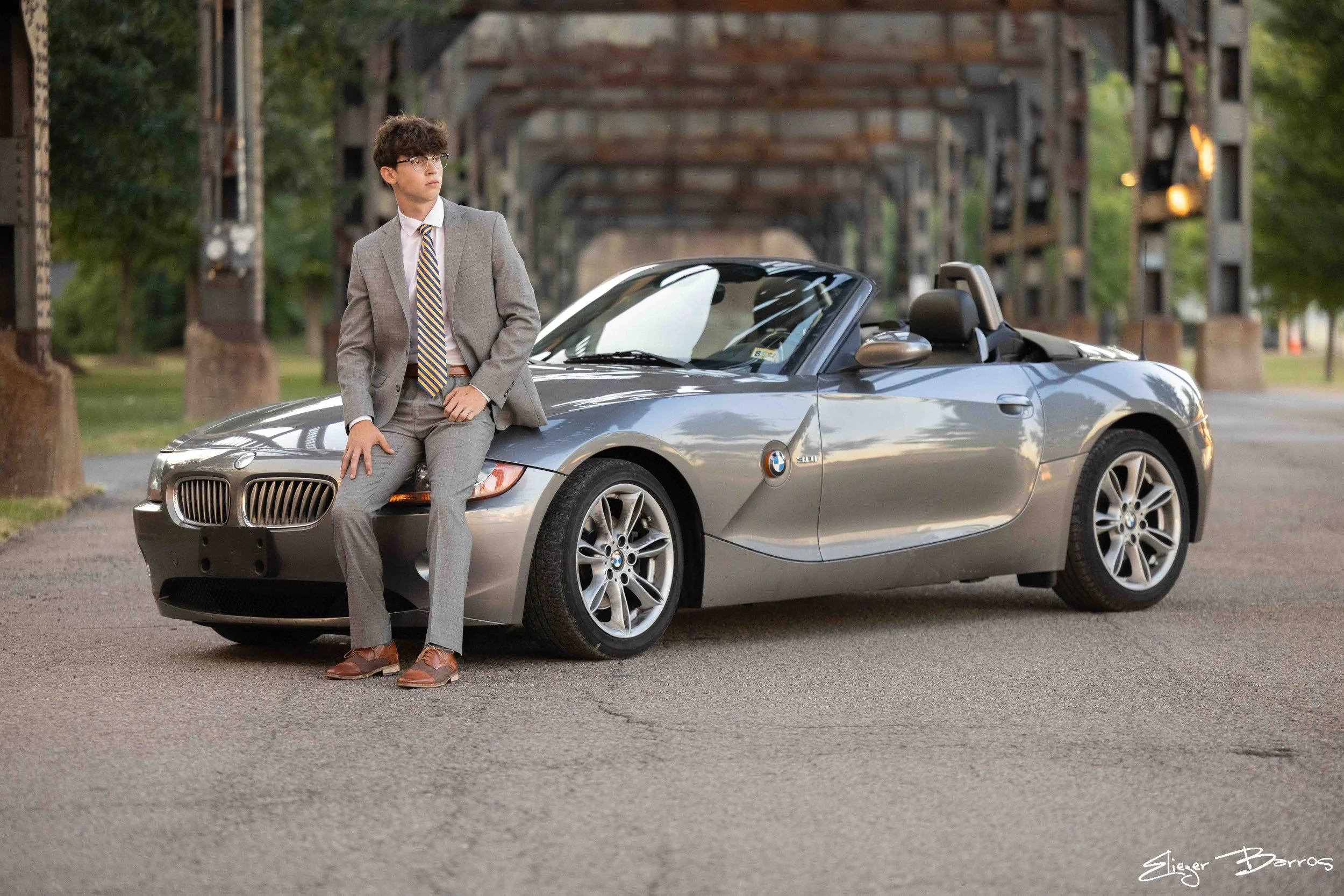 Senior Pictures Pittsburgh in the Strip District. Young man in a gray suit sitting on a silver BMW convertible under a bridge during his stylish outdoor senior photoshoot.
