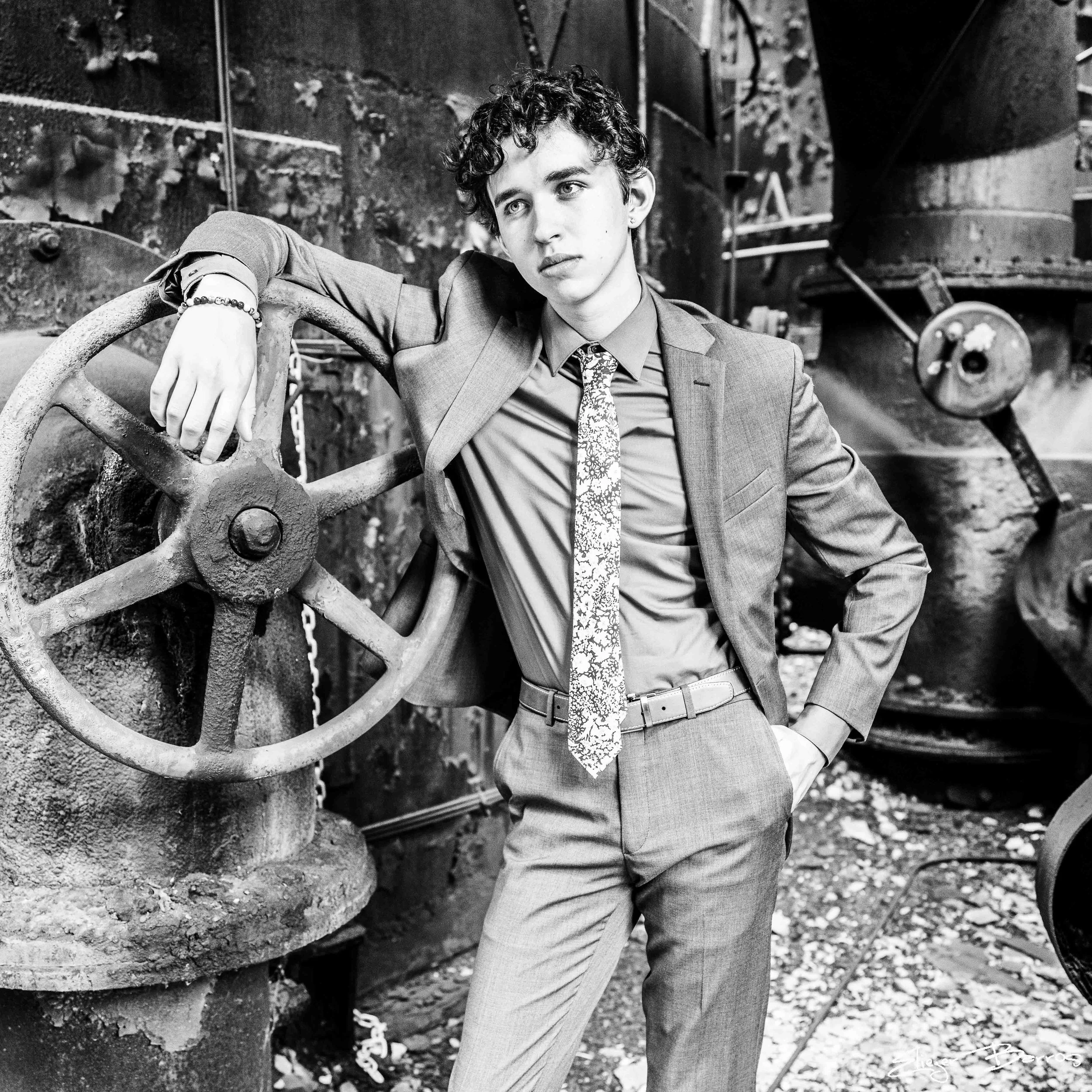 A young man with curly hair dressed in a suit and tie, leaning against a large territorial piece of machinery, in an outdoor industrial setting.