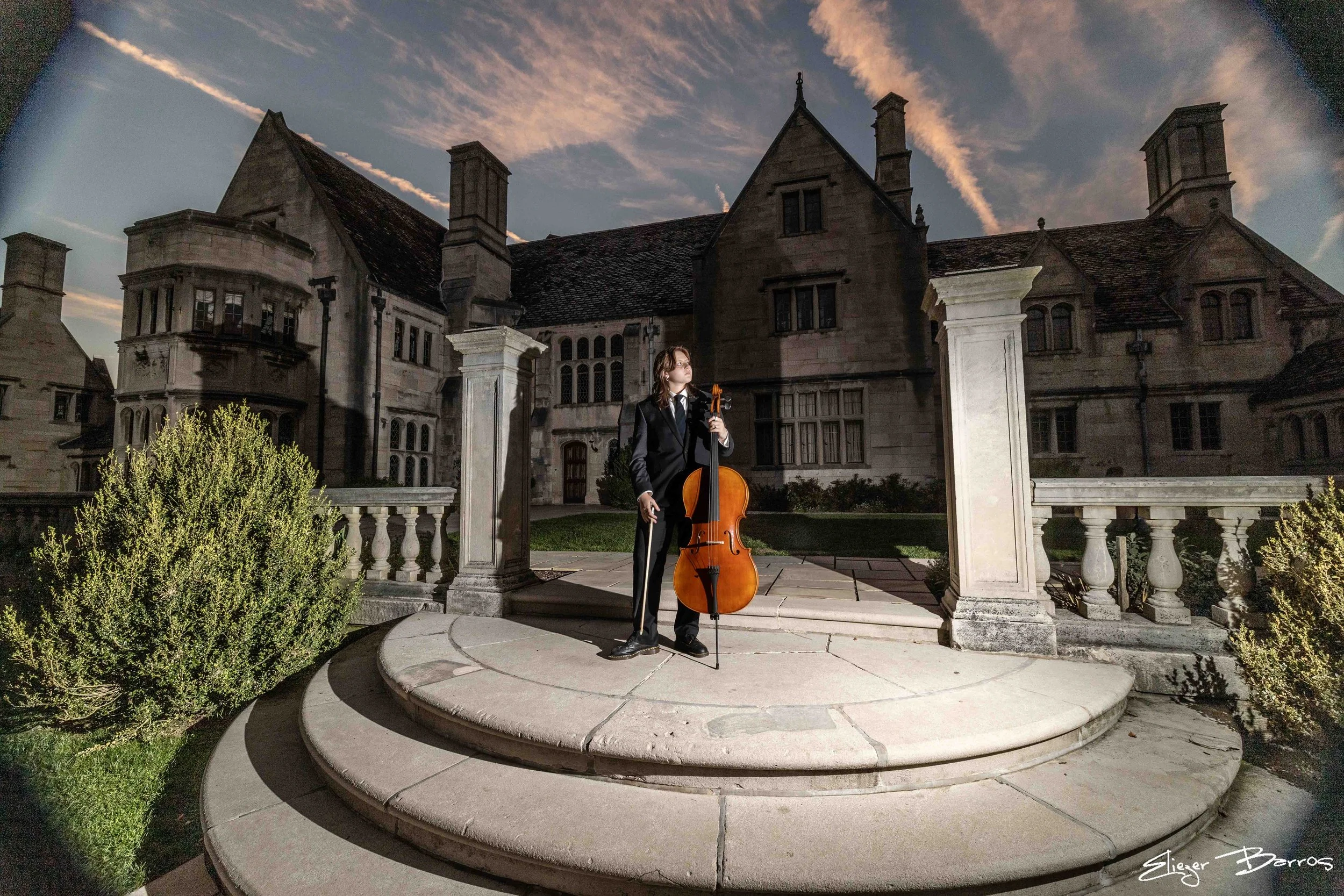 A woman in a black suit playing a cello outdoors on a staircase in front of a large, historic mansion at dusk, with a cloudy sky overhead.