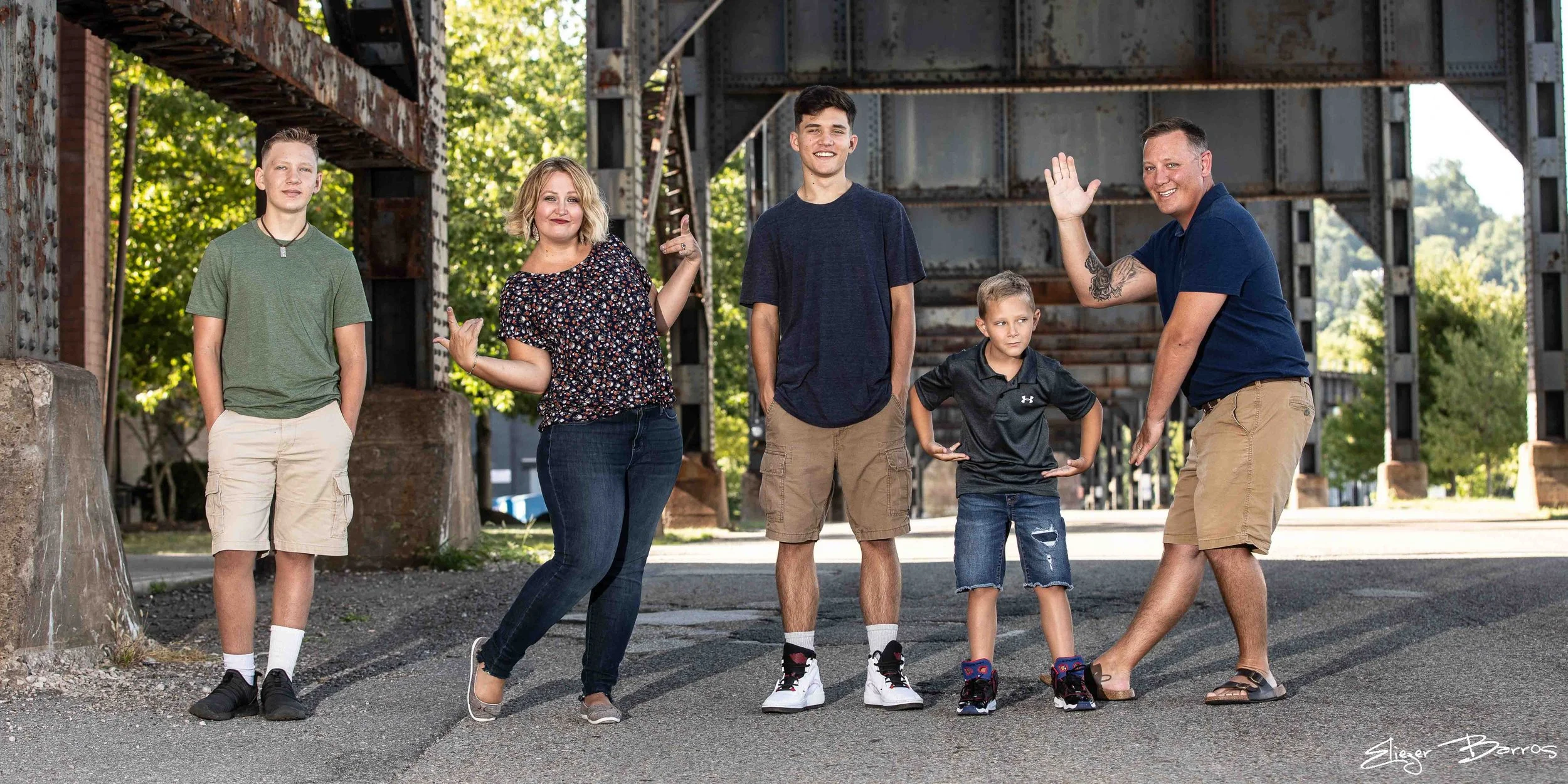 A family of six standing outdoors under a bridge, smiling and waving, with green trees in the background.
