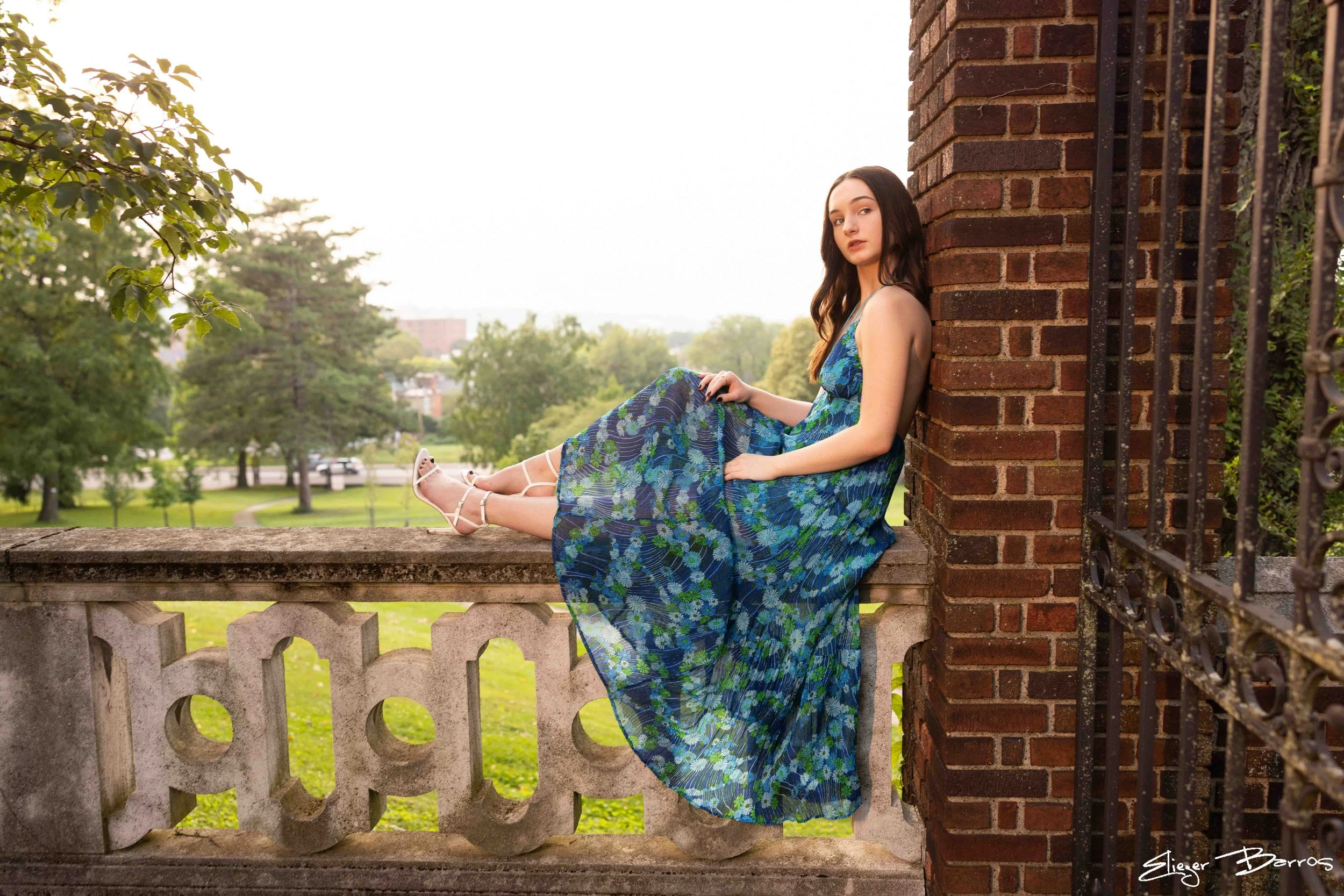 Young woman in a blue floral dress sitting on a stone ledge, leaning against a brick wall, in a park with trees in the background, during sunset.