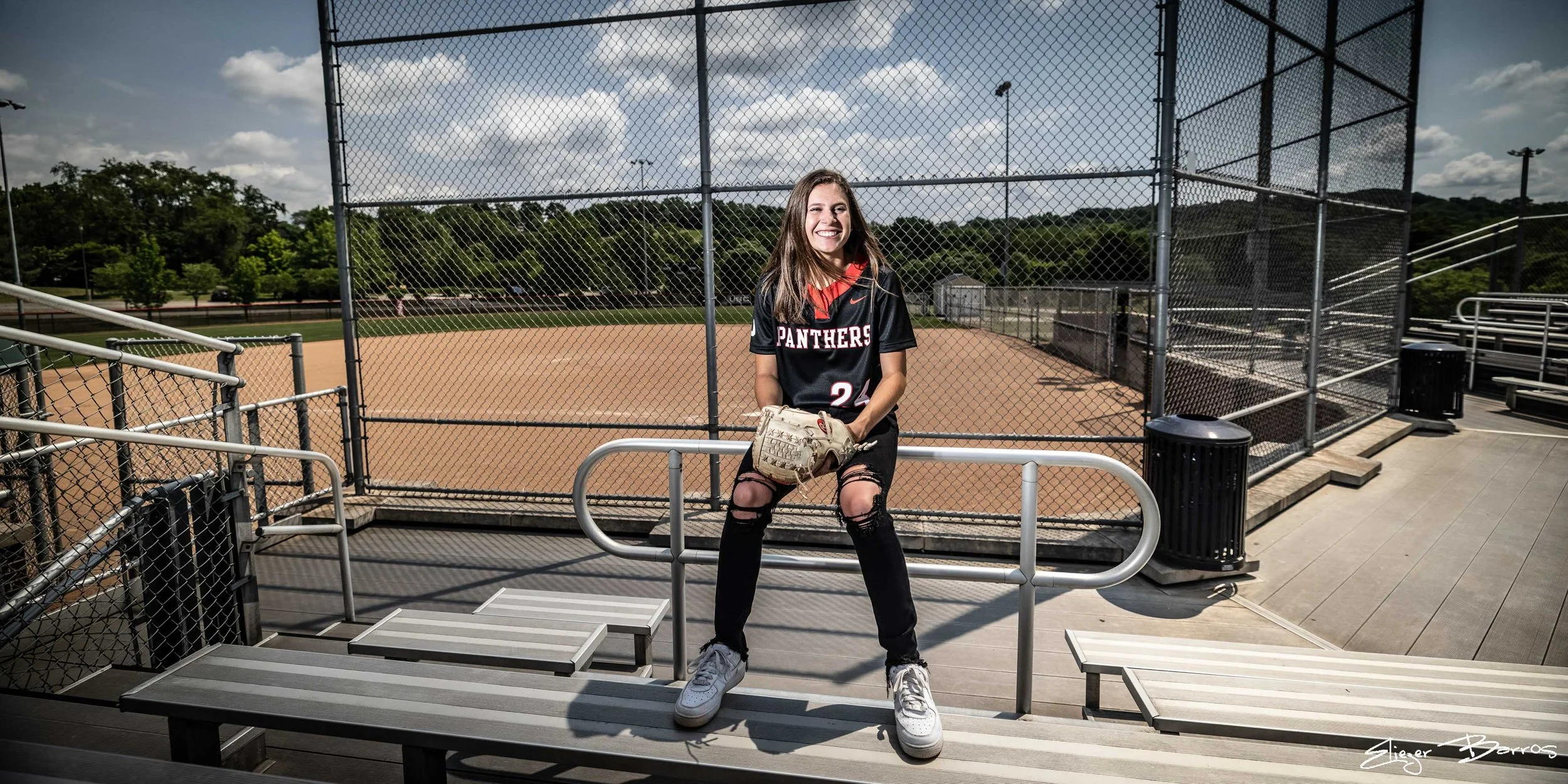Senior Pictures Pittsburgh high school softball. Young woman in Panthers jersey and softball gear posing on bleachers with field in background for athletic senior portraits.