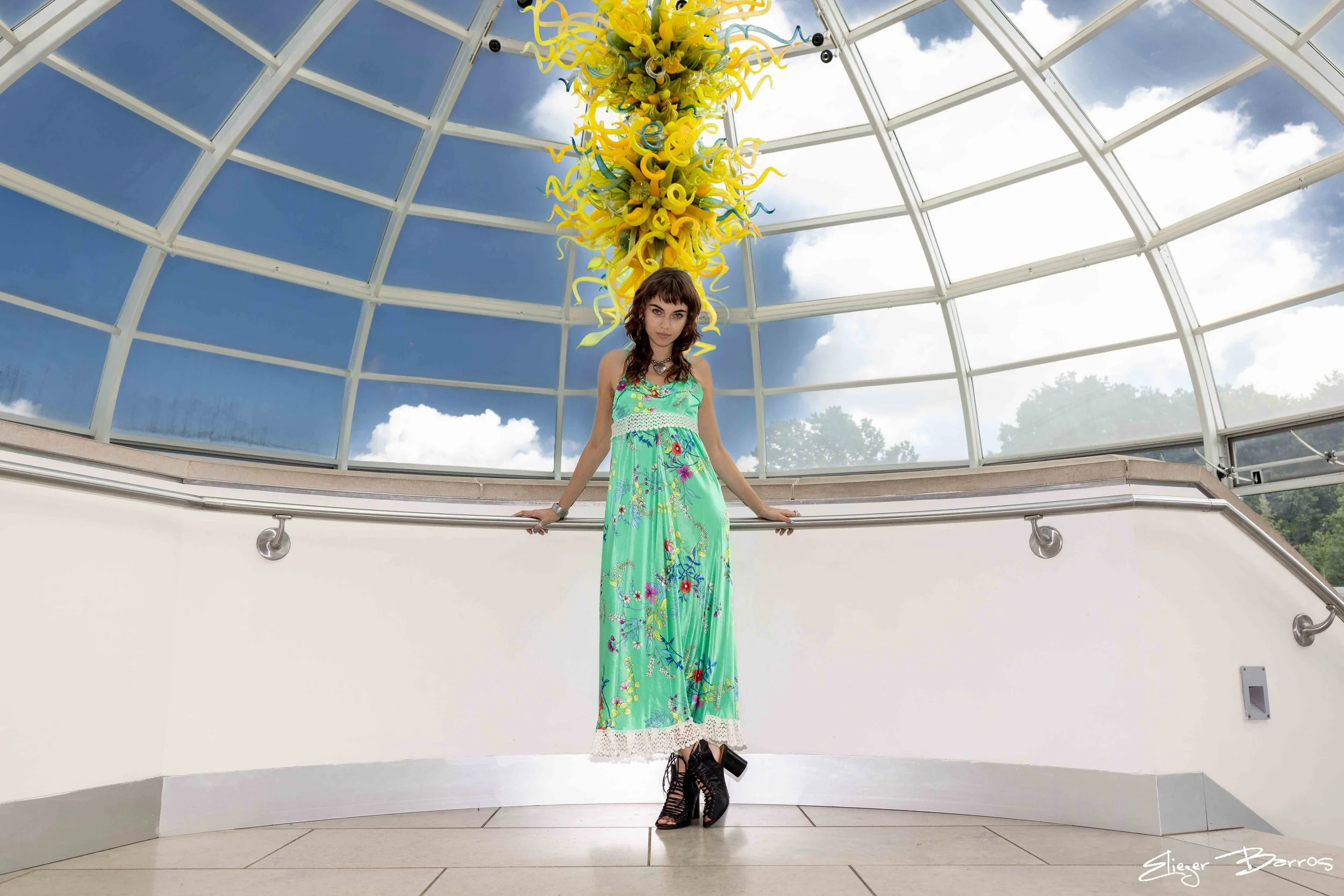 A woman in a long, colorful floral dress and black heeled sandals, standing in an indoor space with a glass ceiling, with a large hanging yellow and blue glass art sculpture above her, and a clear blue sky with some clouds outside