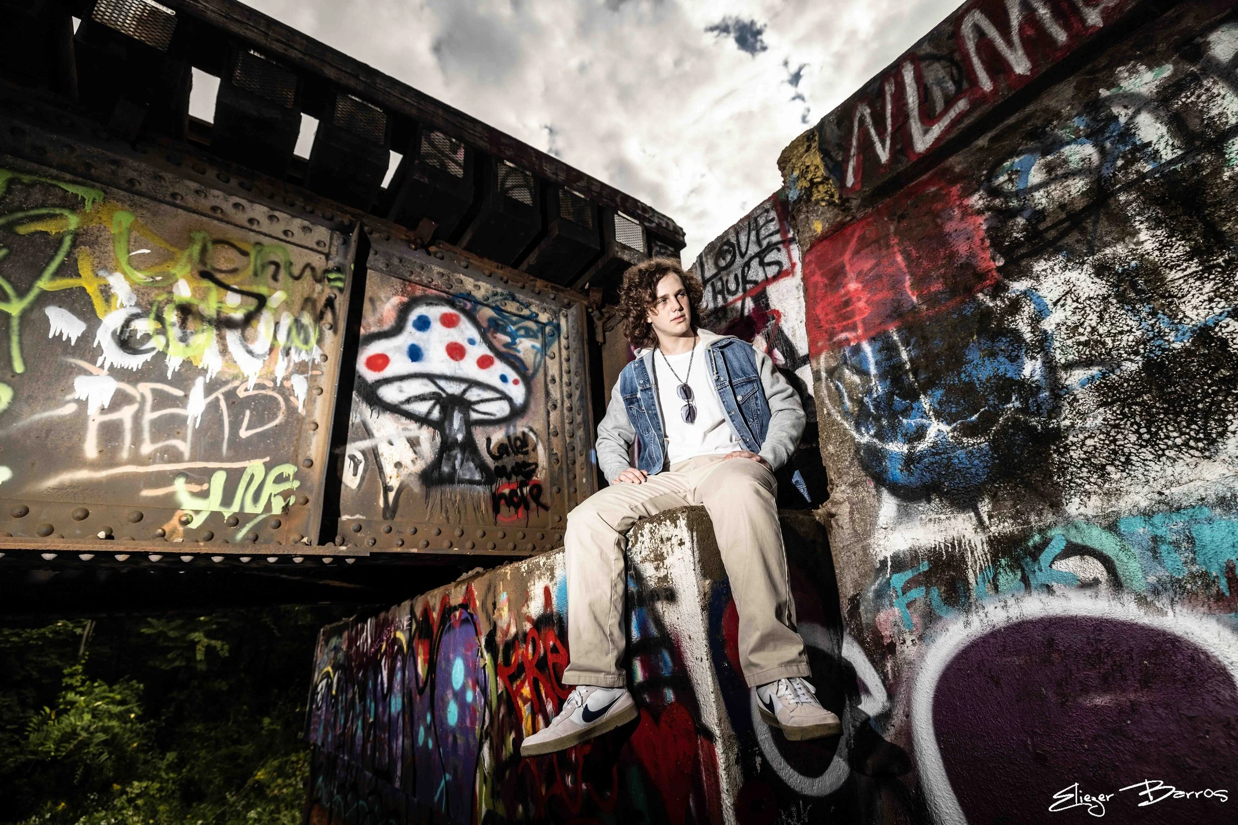 A young man with curly hair in a denim jacket and beige pants sitting on graffiti-covered concrete with a spray-painted mushroom, surrounded by graffiti walls and a cloudy sky.