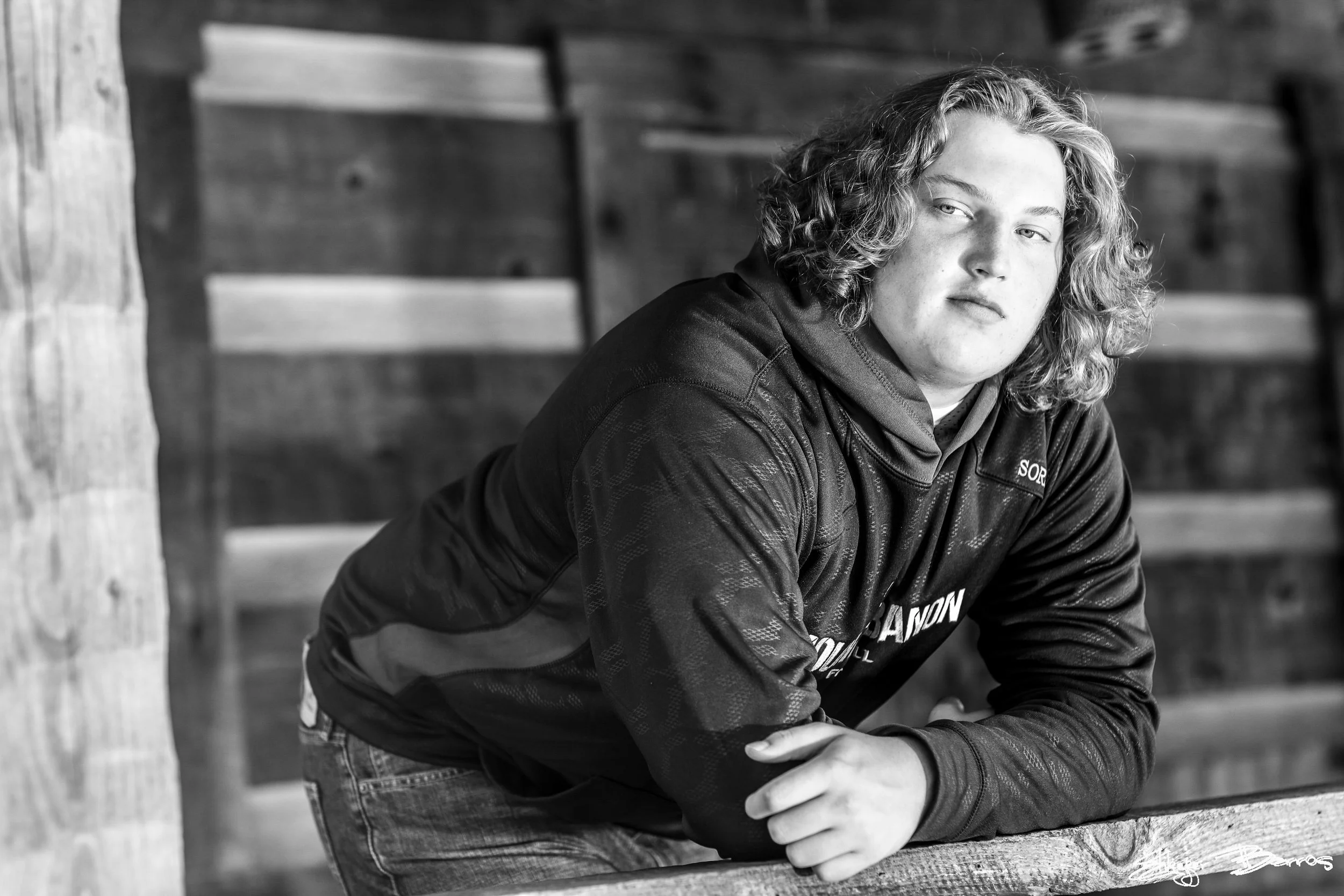 Black and white photo of a young man with curly hair leaning on a wooden railing, with a wooden wall in the background.