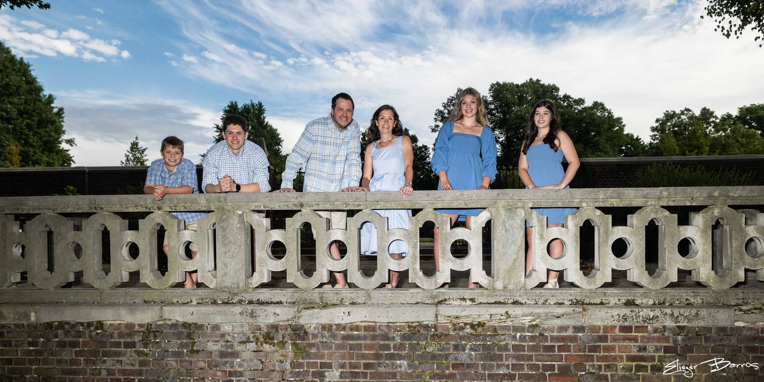 Family of six posing on a stone gazebo with a brick base, trees, and a partly cloudy sky in the background.