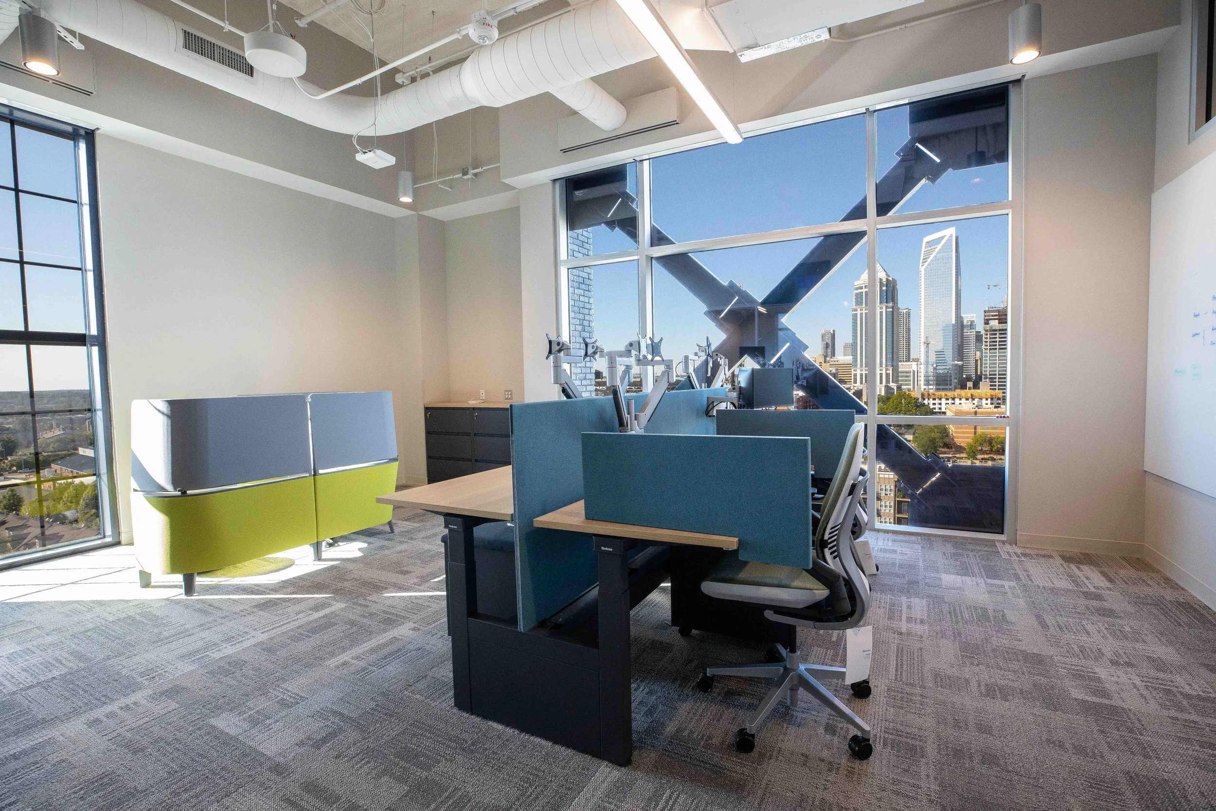 Modern office with large windows overlooking a city skyline, featuring a spiral staircase outside and designed workspaces with chairs and desks.