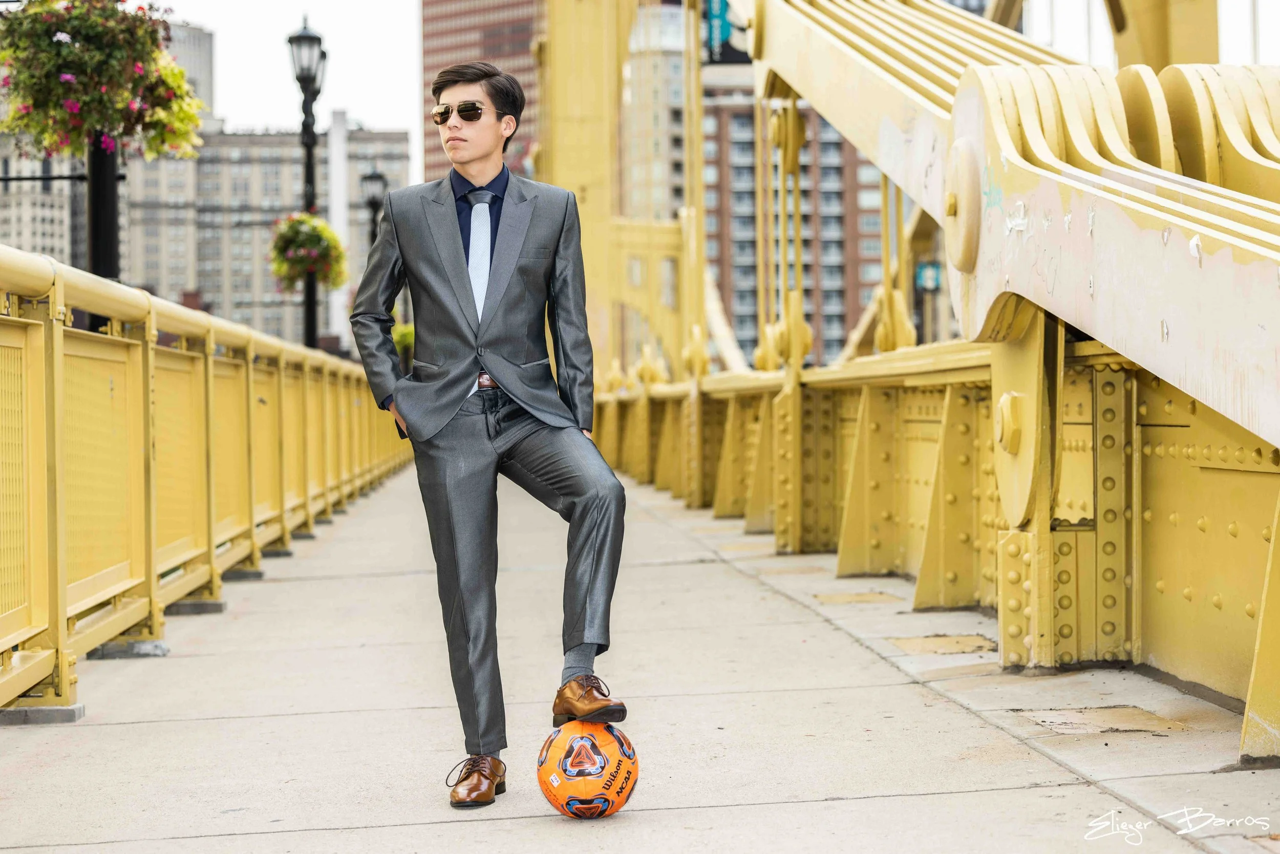 A young man in a gray suit, sunglasses, and brown shoes standing on a bridge, with one foot on an orange and blue soccer ball, in an urban setting with yellow safety barriers and city buildings in the background.