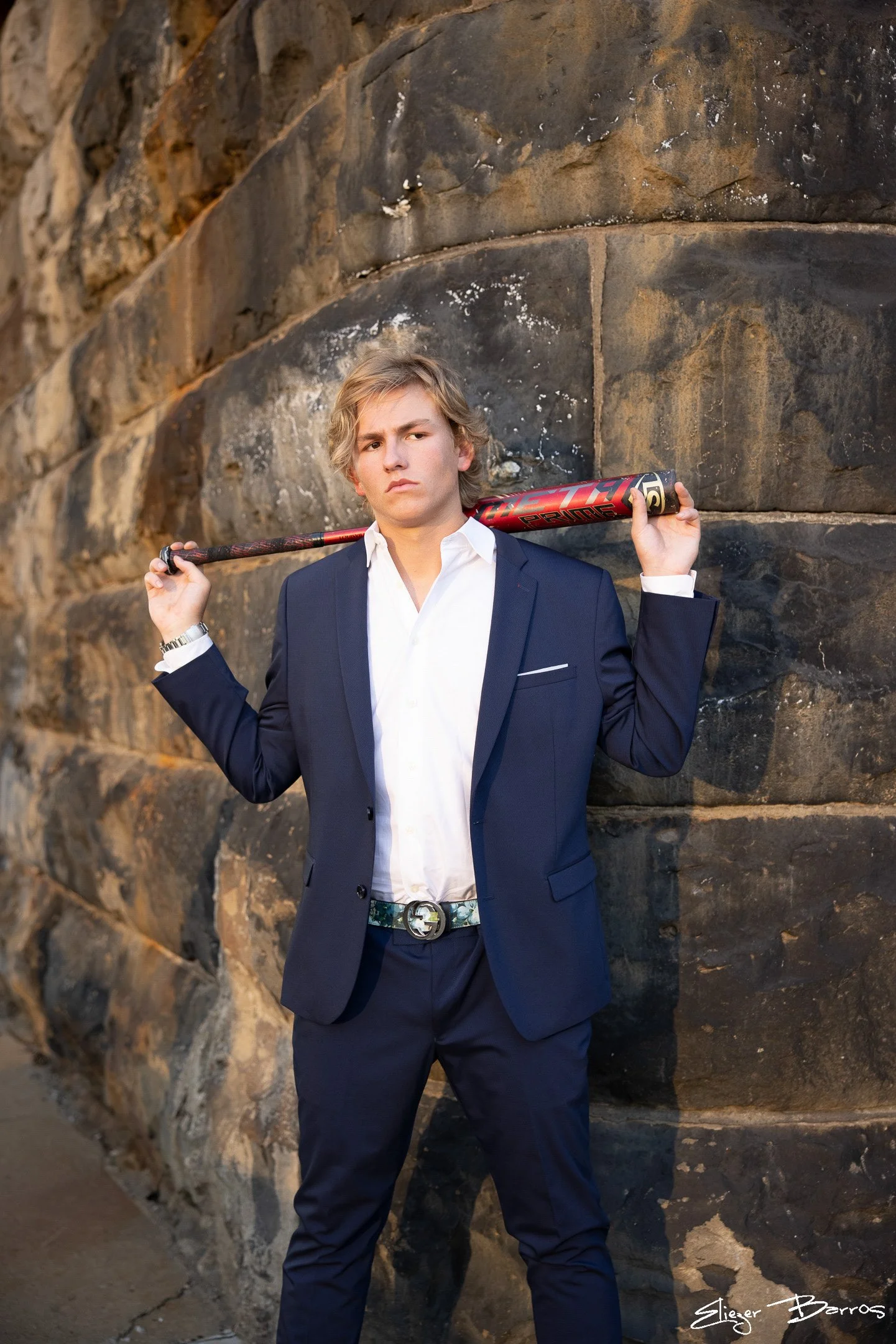 Young man in a navy suit and white shirt standing against a large stone wall, holding a baseball bat over his shoulder.