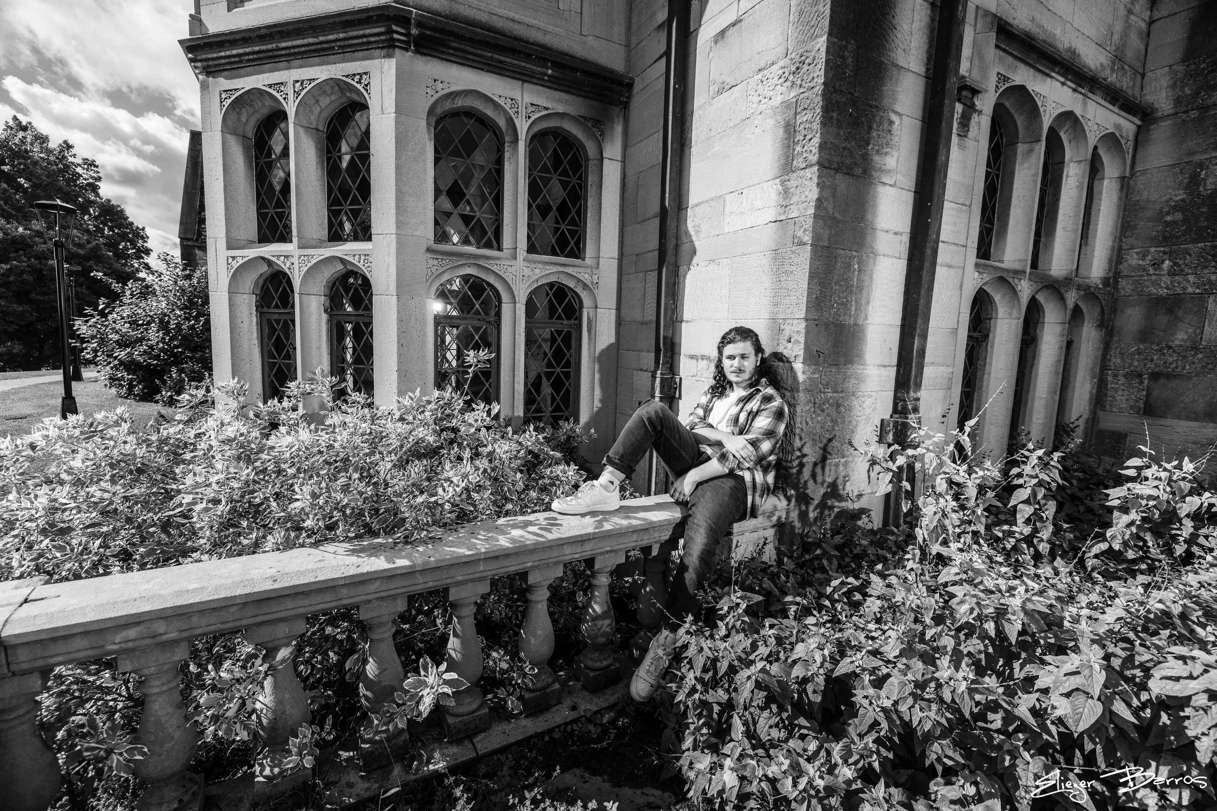 A young person with long hair sitting on a stone ledge in front of a historic building with arched windows. The scene is captured in black and white, with lush bushes in the foreground and a partly cloudy sky in the background.