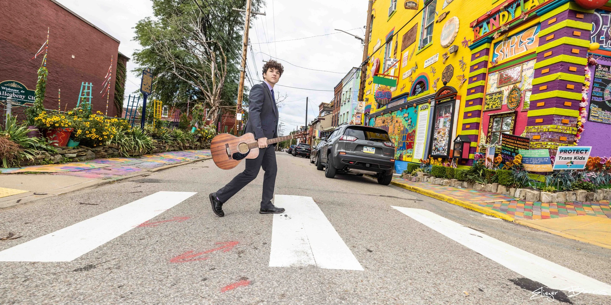 A man in a dark suit walking across a pedestrian crosswalk holding an acoustic guitar in a colorful urban street scene. There are vibrant murals, signs, cars, flowers, and trees along the street.