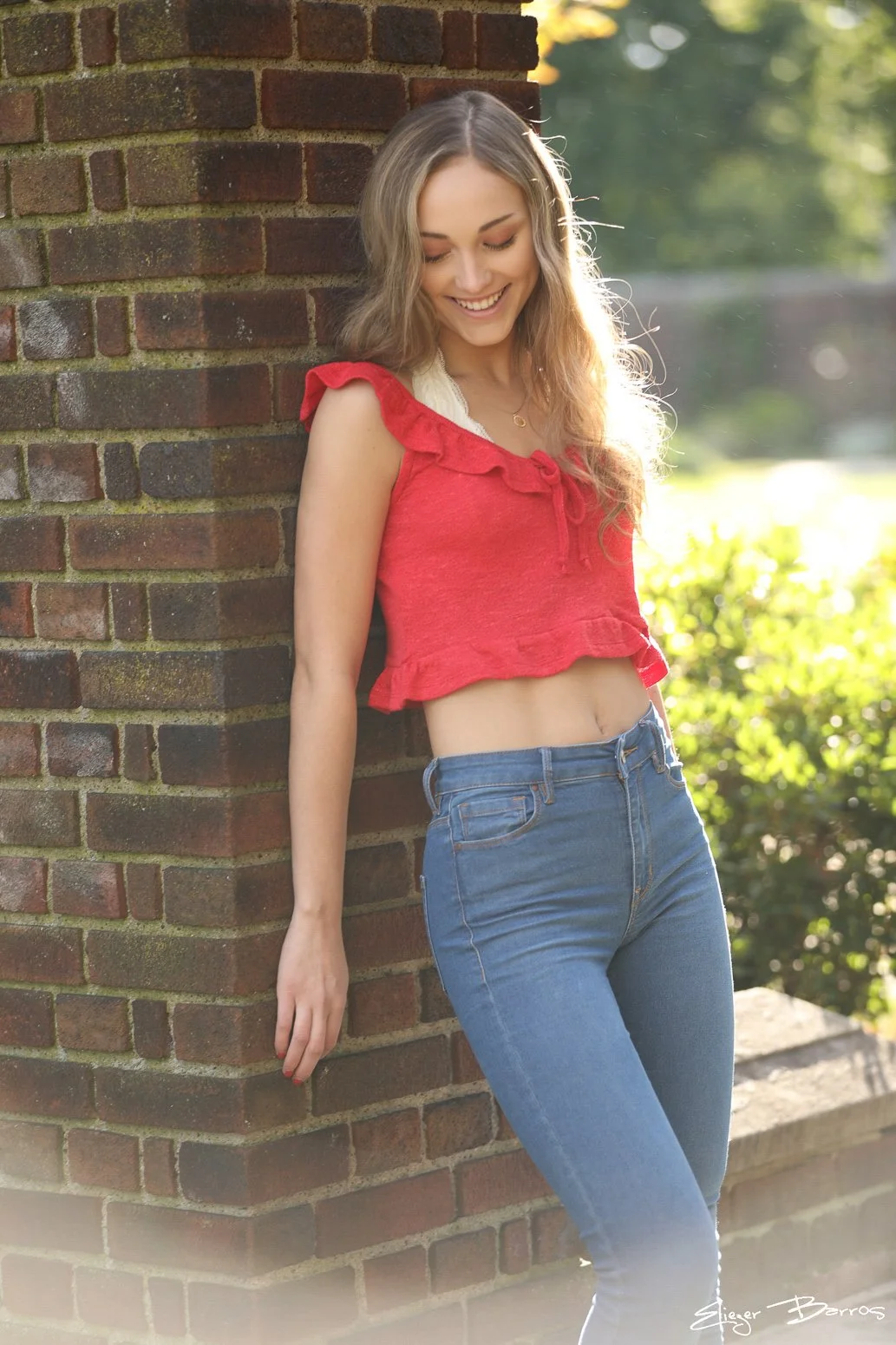 A young woman with long wavy hair, wearing a red sleeveless crop top with ruffles and blue jeans, is standing outdoors leaning against a brick wall, smiling with her eyes closed in bright sunlight.