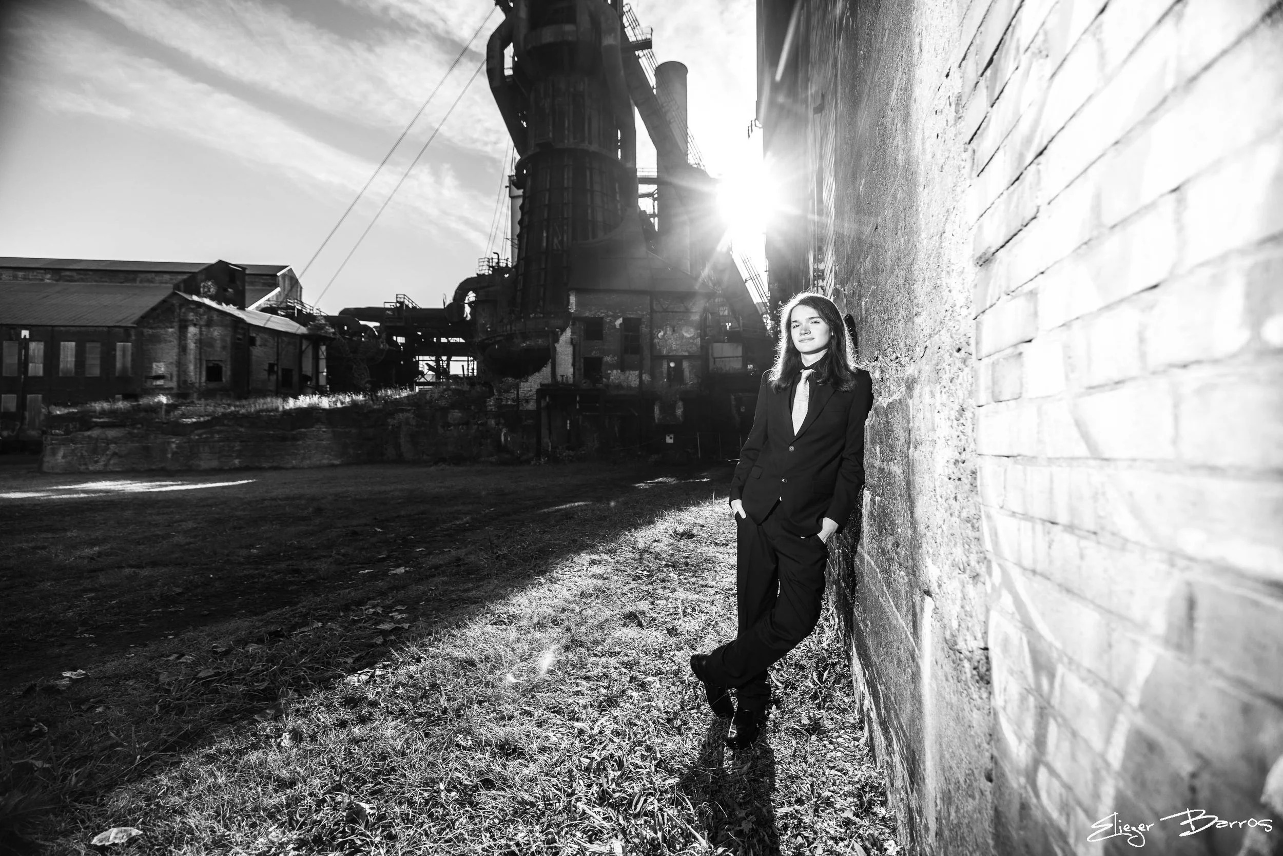 A young woman in a suit standing outdoors against a brick wall, with an industrial factory and cloudy sky in the background. The sun is shining behind her, creating a silhouette effect.