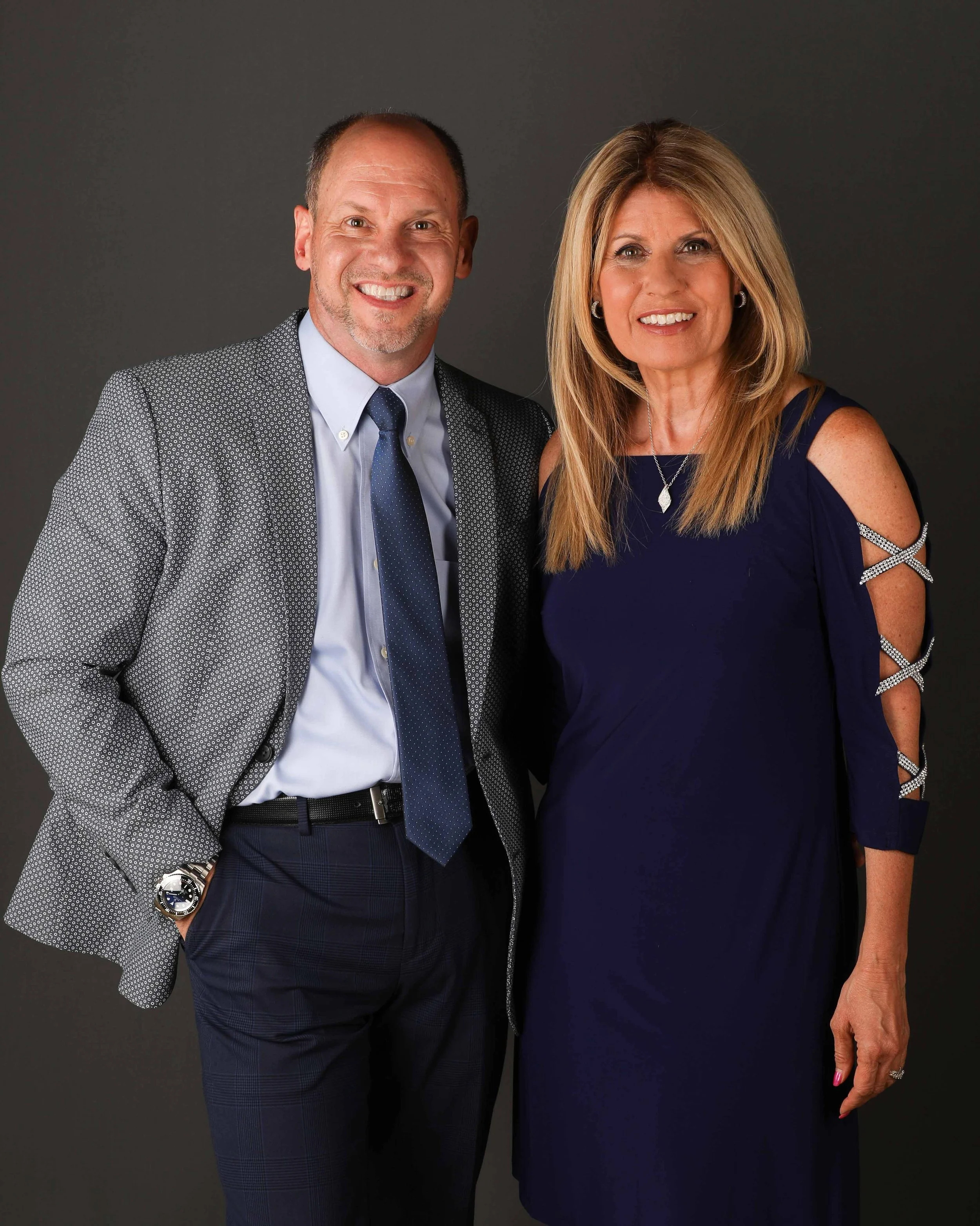 A man in a gray patterned blazer with a light blue shirt and navy tie stands next to a woman in a dark blue dress with cut-out shoulder details and diamond jewelry, both smiling against a dark background.