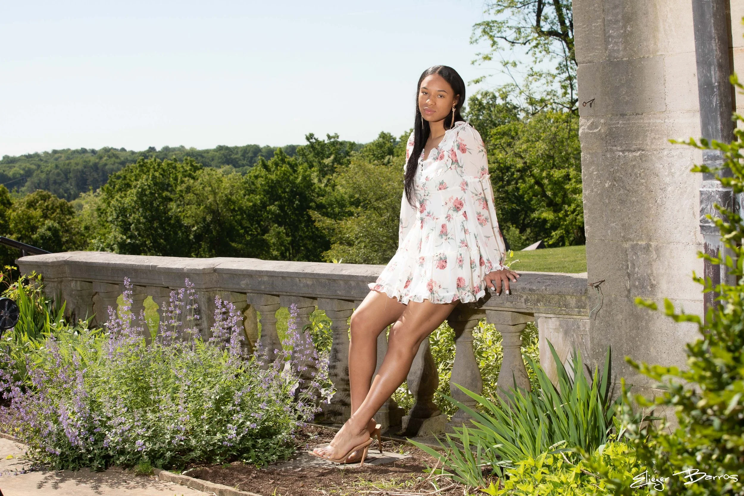 A young woman in a floral dress and high heels sitting on a stone railing in a garden with trees and flowering plants, on a sunny day.