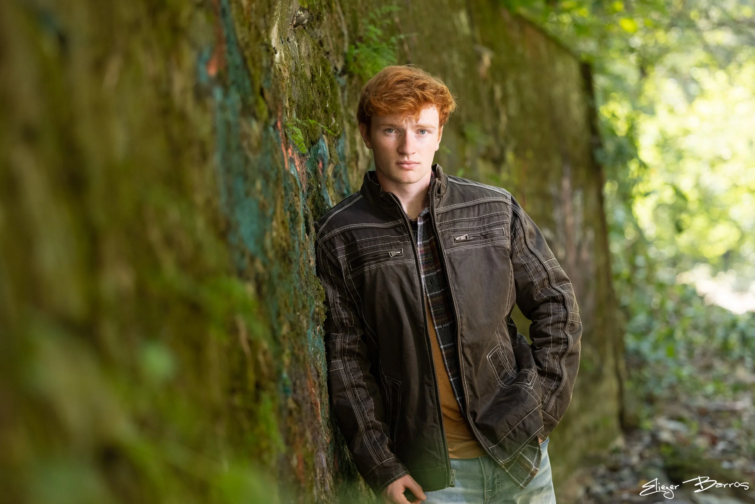 Young man with red hair wearing a black jacket standing outdoors beside a moss-covered brick wall in a wooded area
